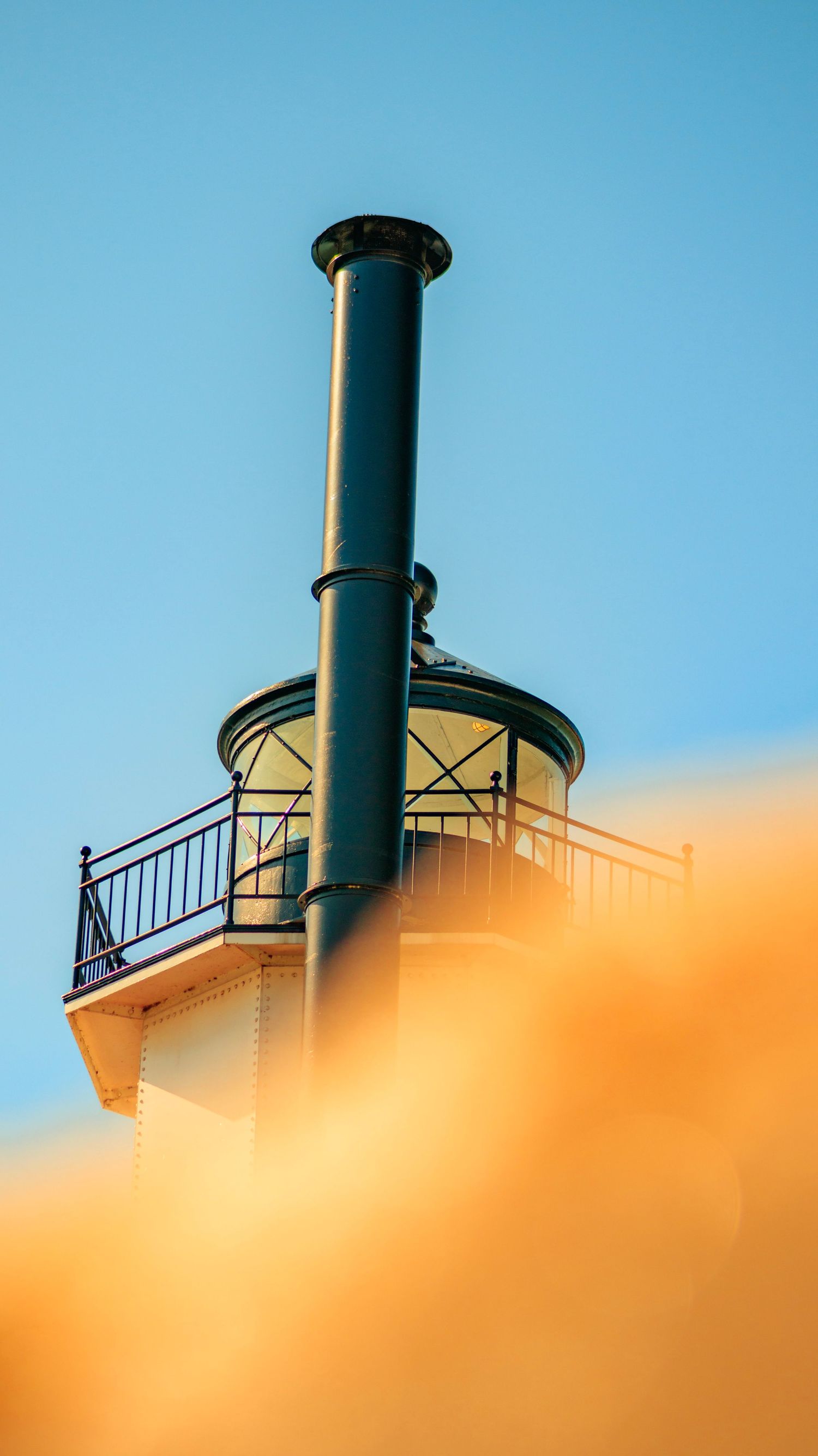 Artistic close-up of lighthouse tower with railing against orange and blue sky gradient.