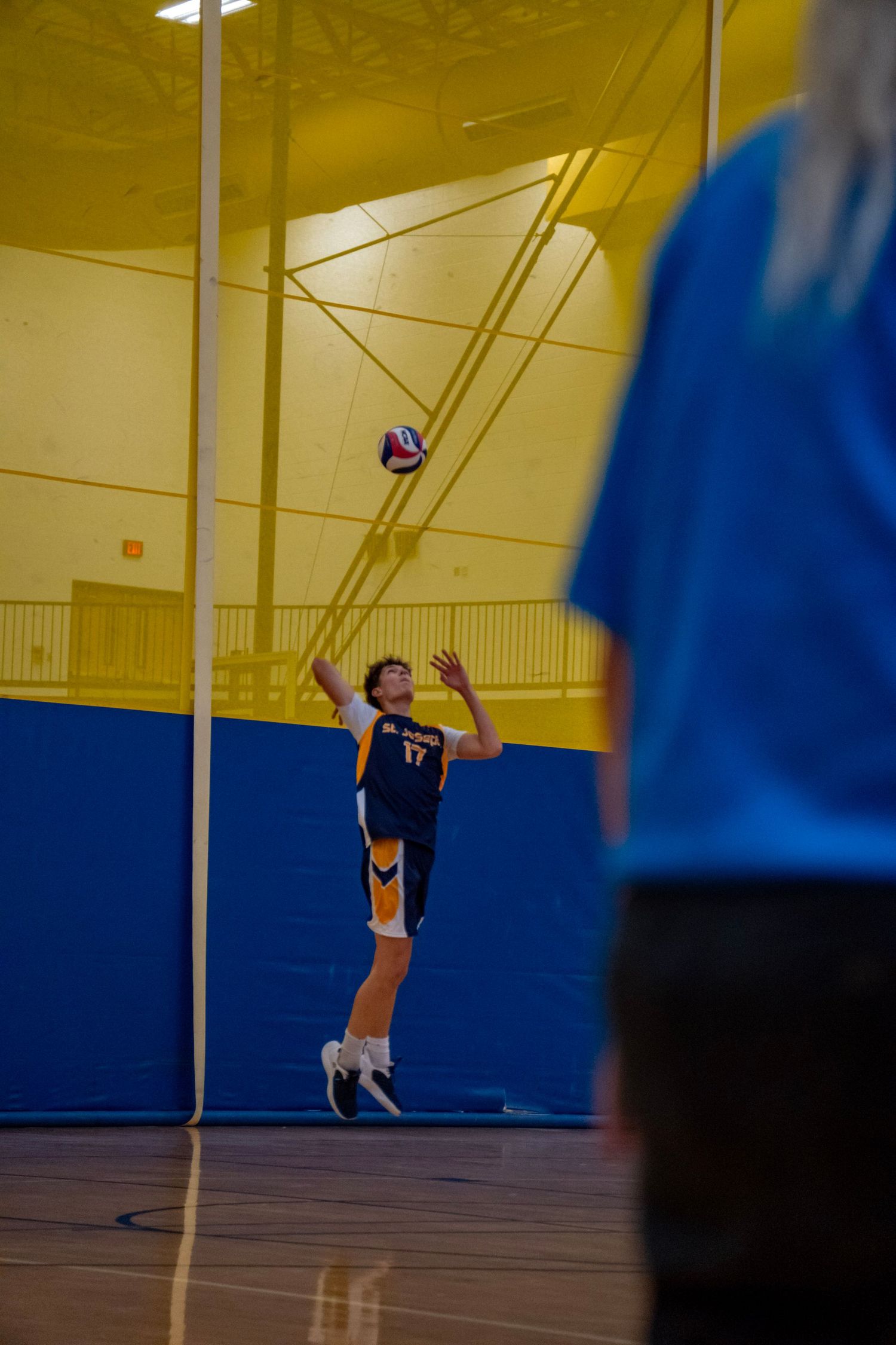 Volleyball player jumping to serve the ball in indoor gymnasium with blue and yellow walls.