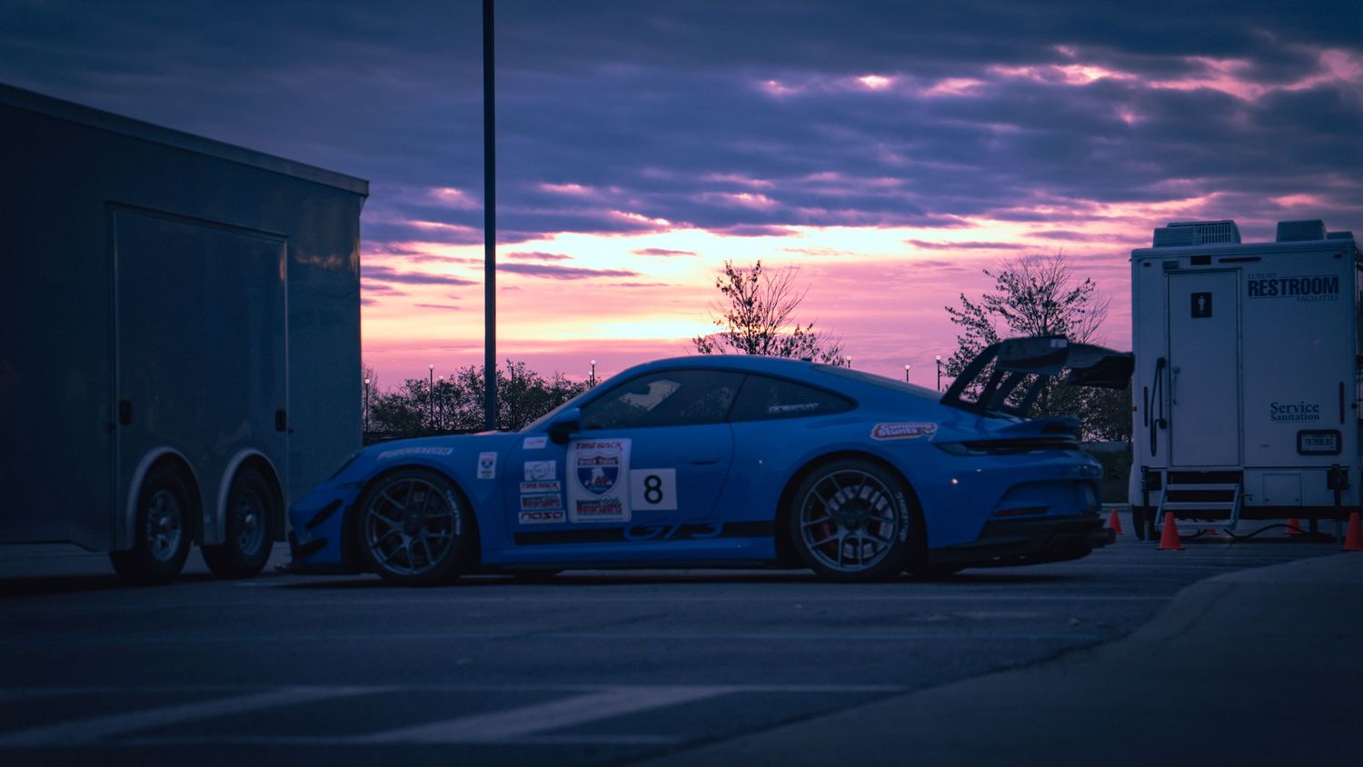 Blue racing Porsche parked between trailers during colorful sunset.