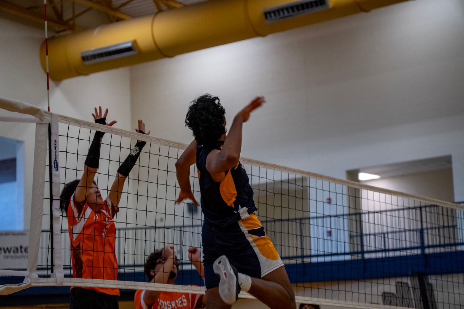 Players jump at the net during a competitive indoor volleyball match.