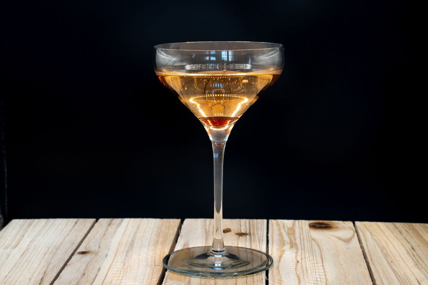Amber cocktail in crystal coupe glass photographed on light wooden surface with dramatic lighting.