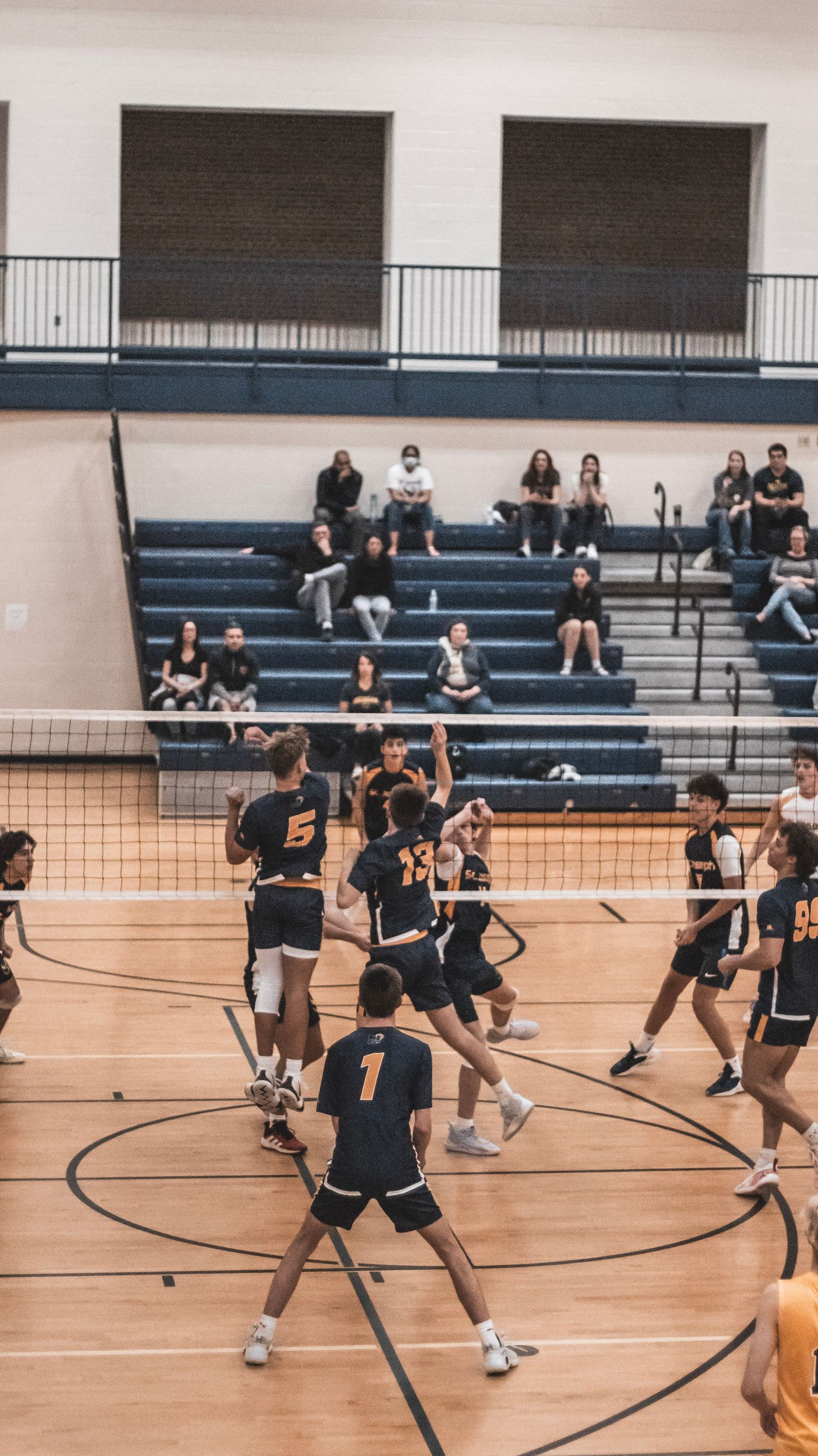 Indoor volleyball match in progress with teams in black uniforms and spectators watching from bleachers.