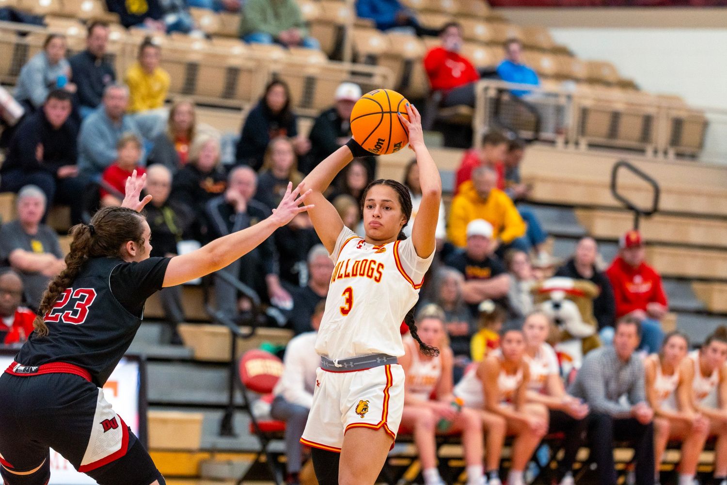 Basketball players compete for possession during an intense game with spectators watching from the stands.