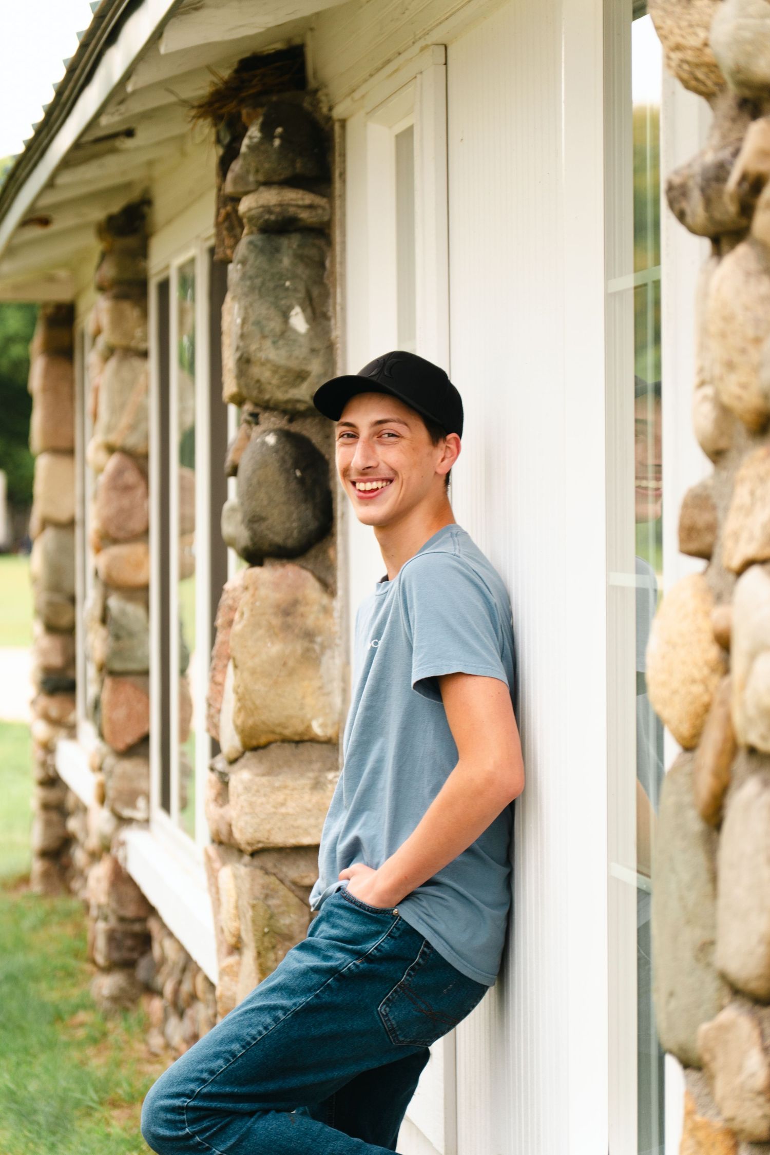 Smiling young man in a black cap, blue t-shirt and jeans leans against a white wall beside a rustic stone pillar and windows.
