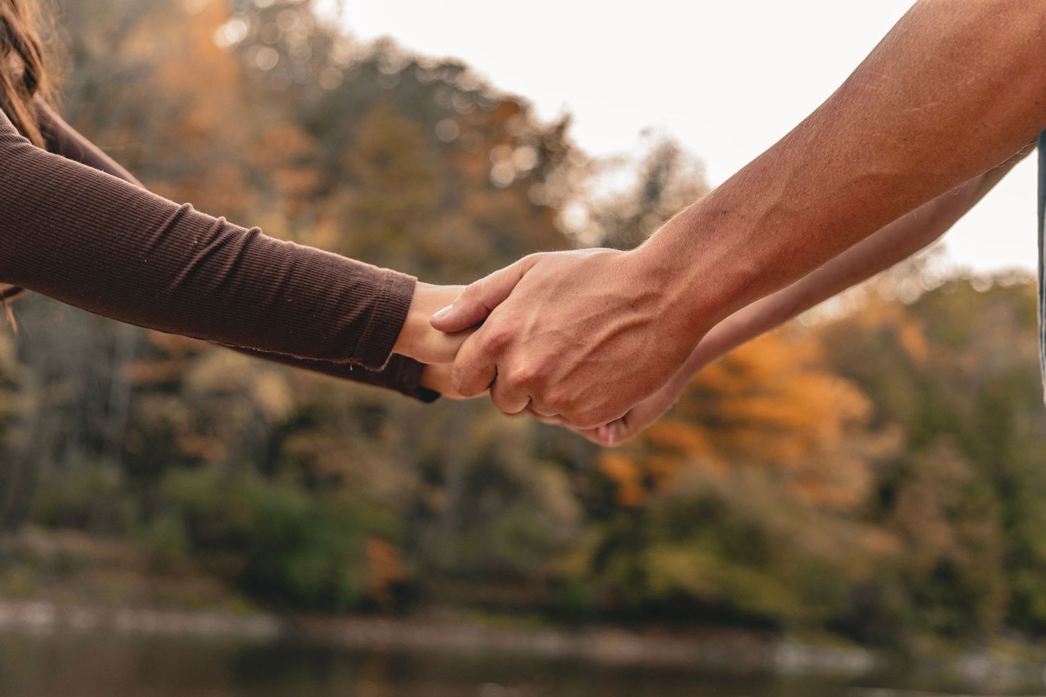 Two people holding hands outdoors beside a calm lake, with colorful autumn foliage softly blurred in the background.