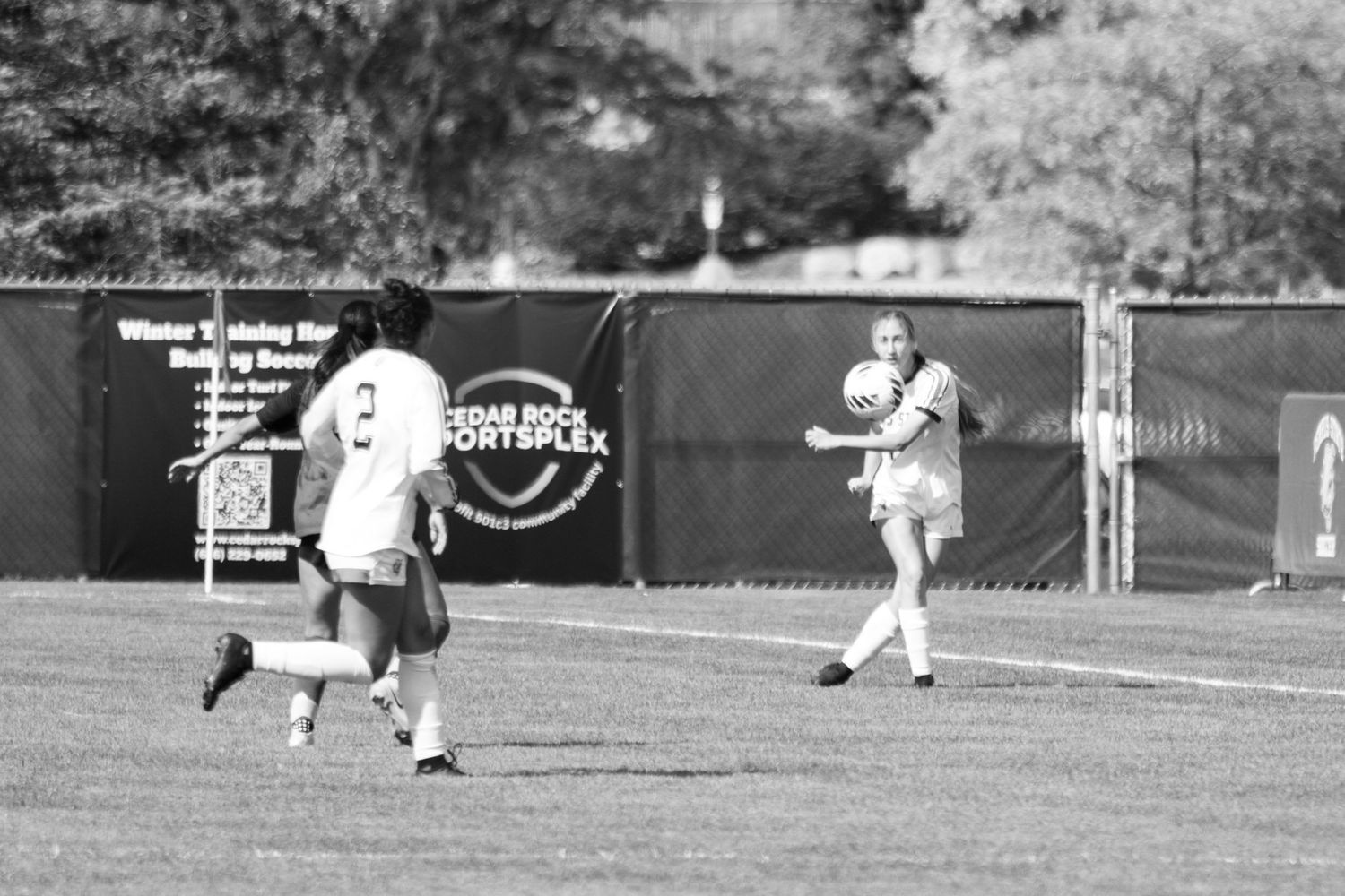 Black and white photo of two women's soccer players on a grass field near a Cedar Bend Sportsplex banner during a match.