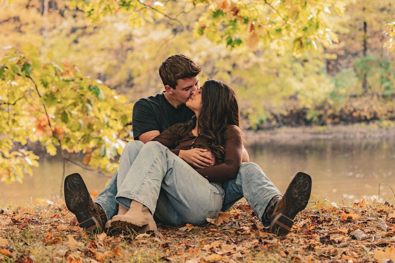 A couple sits close and kisses on a bed of fallen autumn leaves beside a calm lake, surrounded by golden fall foliage.