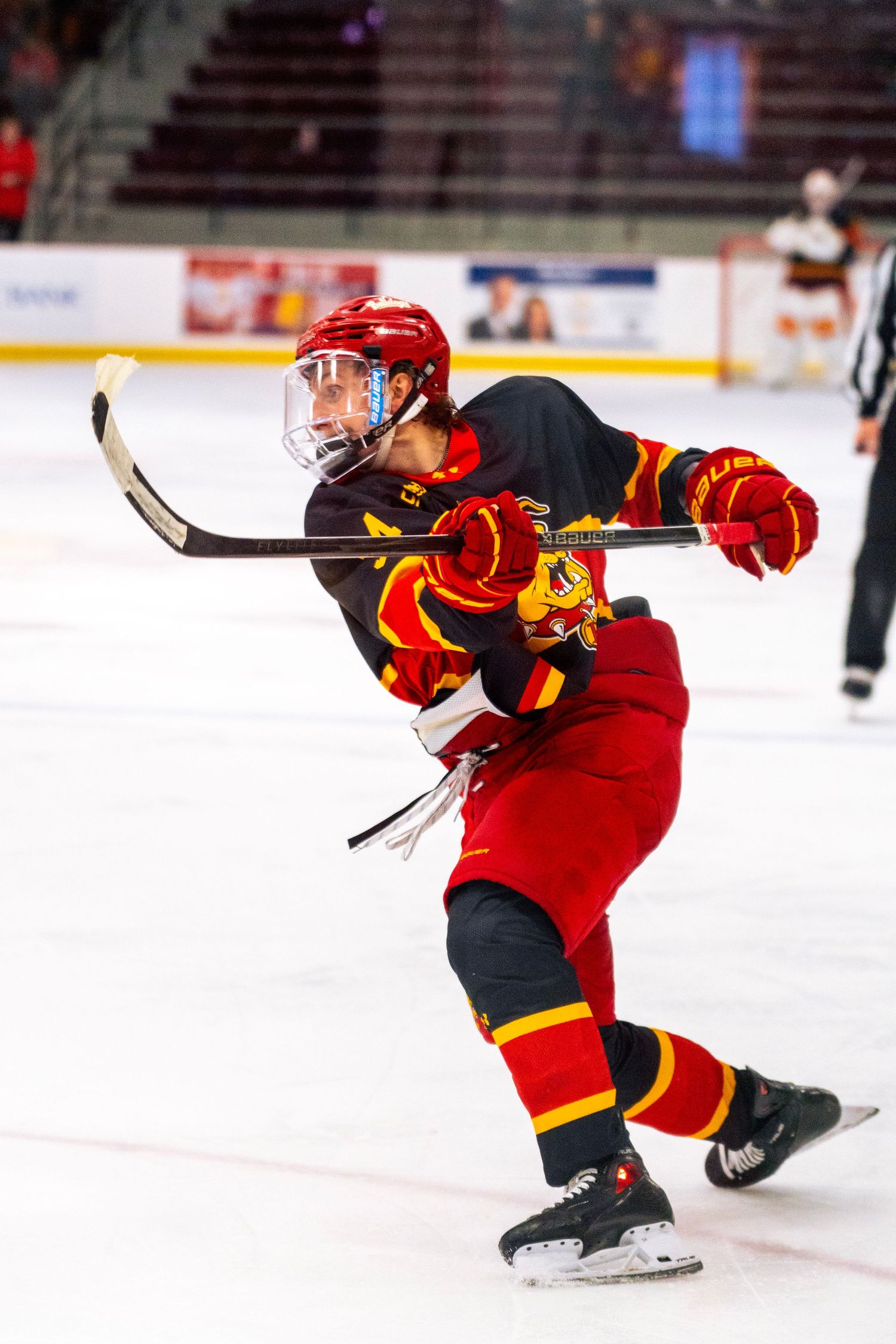 Ice hockey player in a red helmet with visor and red and gold uniform follows through on a shot while skating on the rink.