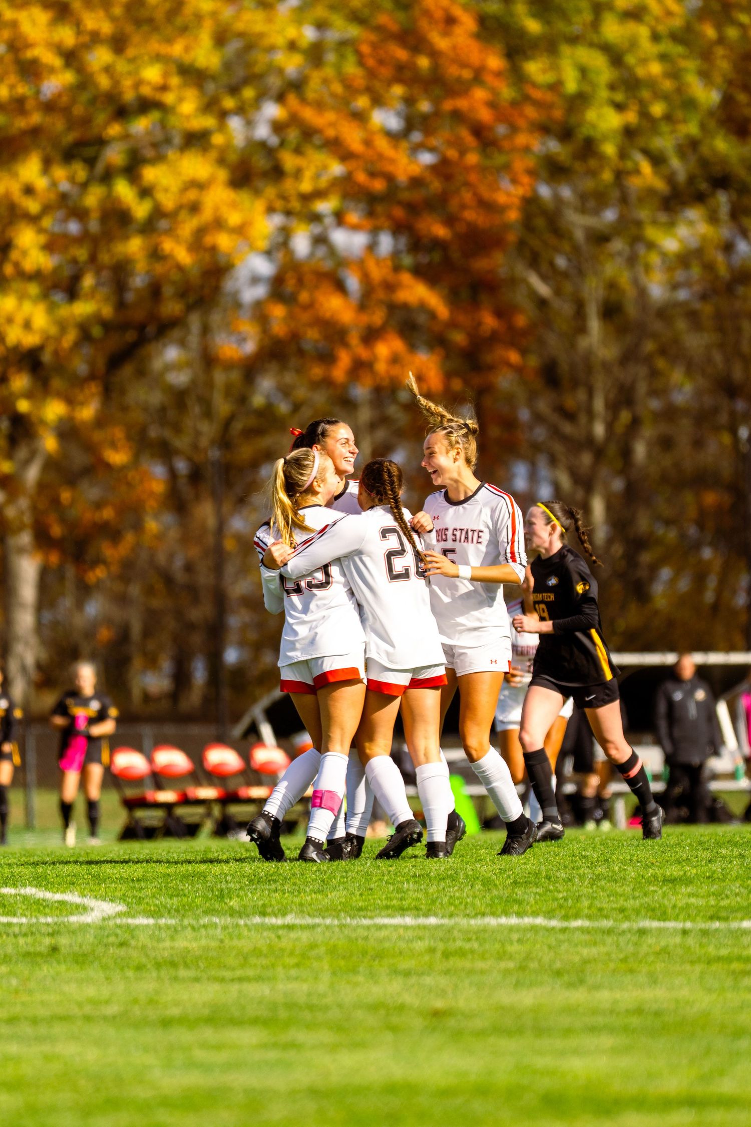 Women's soccer players in white and red uniforms celebrate together on a grass field with colorful autumn trees in the background.