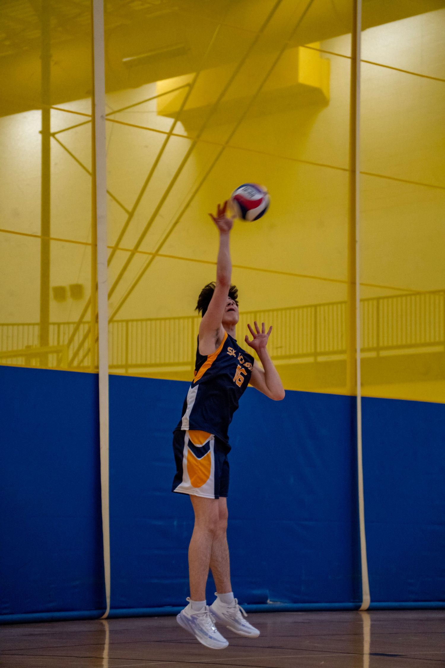 Volleyball player in navy and gold uniform leaps to serve ball during indoor game.