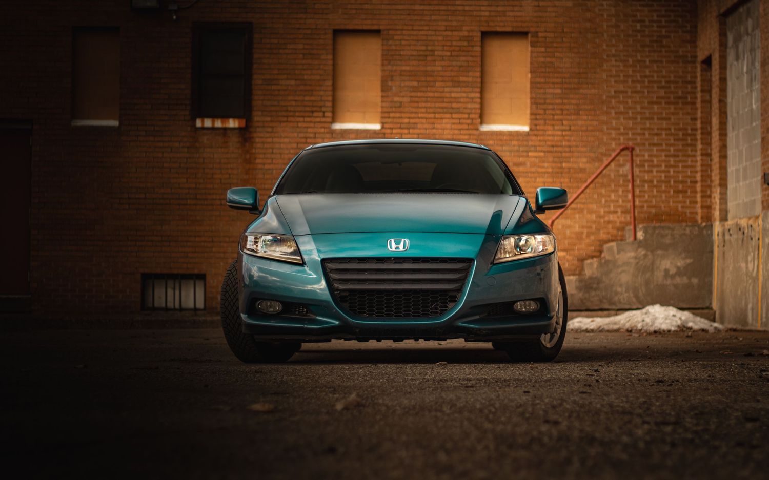 Teal Honda CR-Z sport hybrid car parked in front of brick building at sunset.