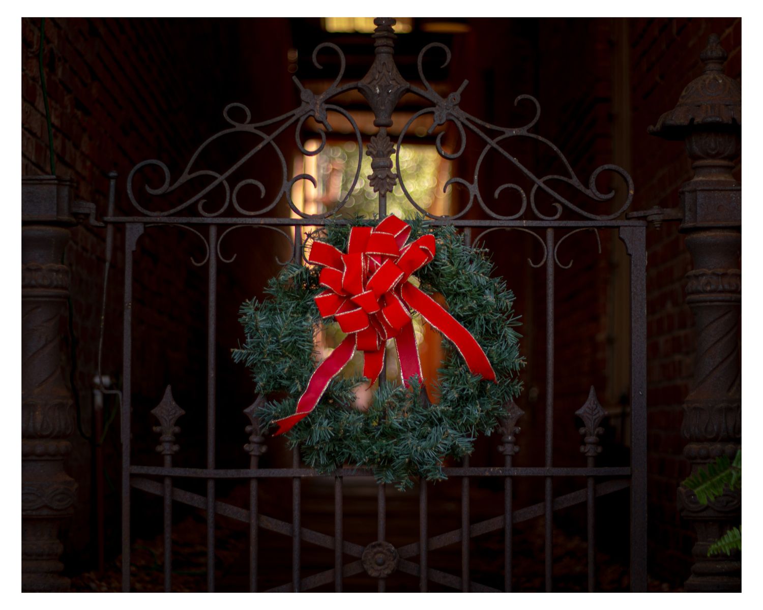 Illuminated Christmas wreath with red bow hanging on decorative iron gate.