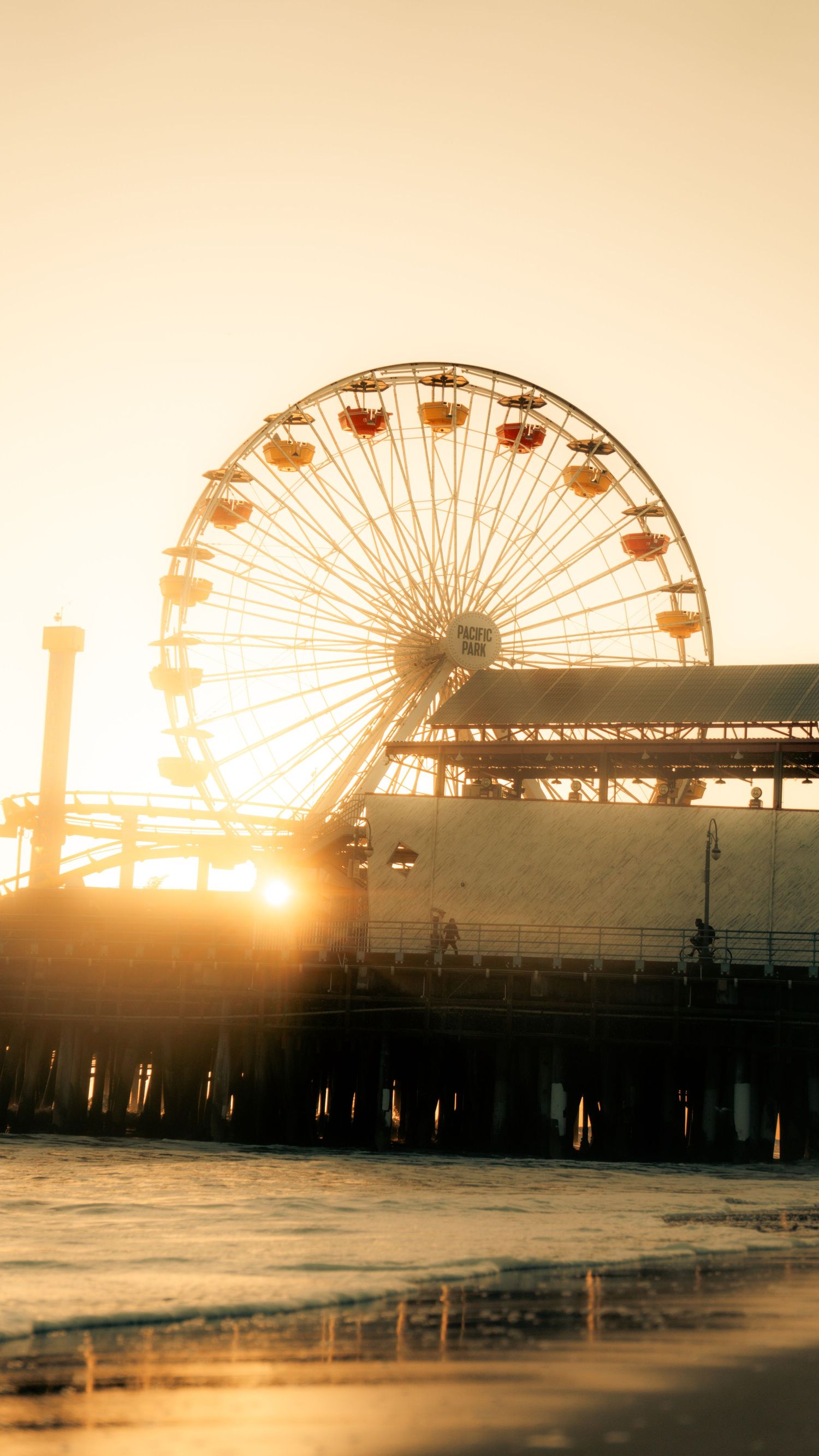 Low angle view of Ferris wheel silhouetted against sunset with waves in foreground.