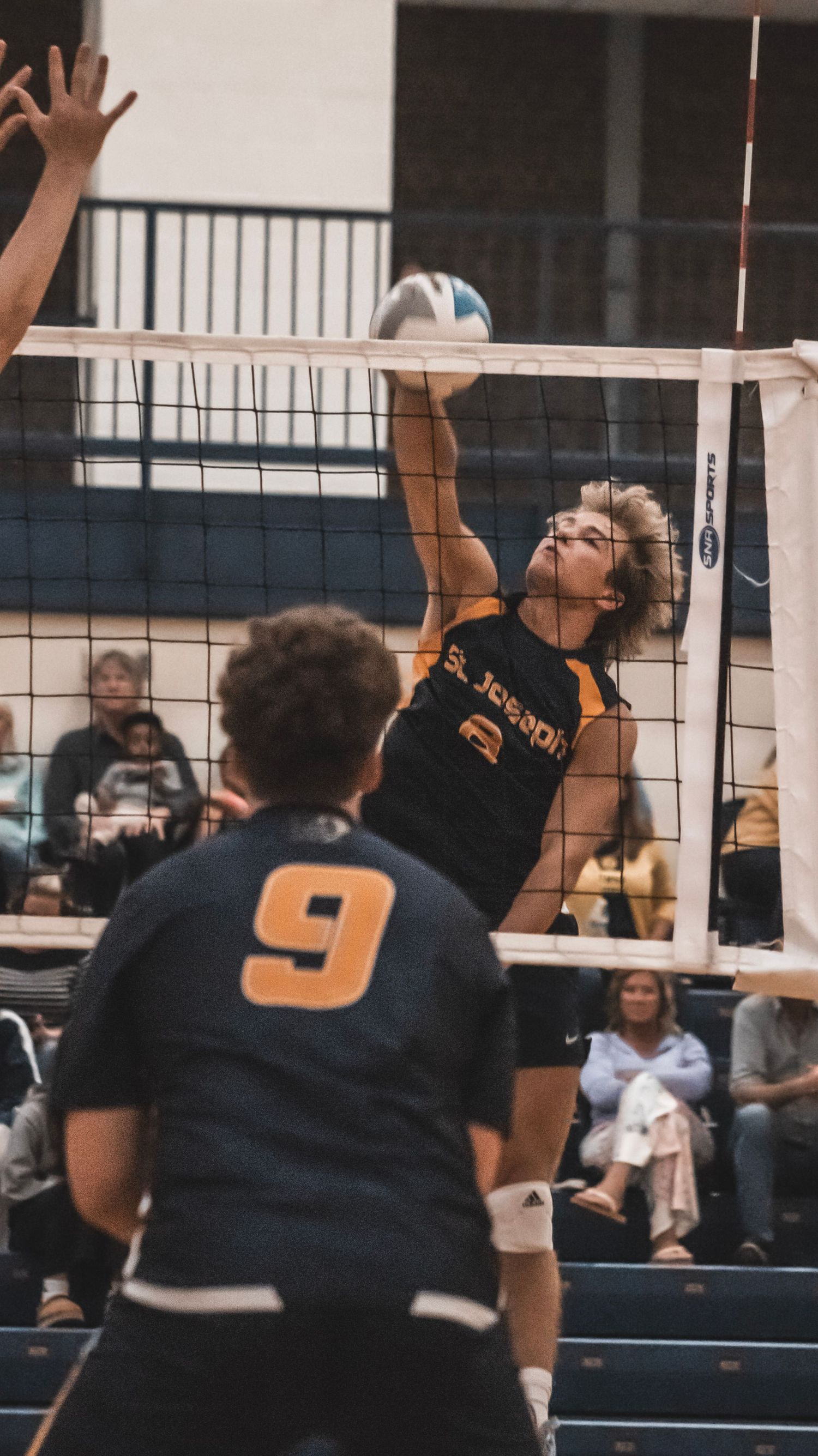 Player leaps high to spike volleyball over net during intense indoor match.