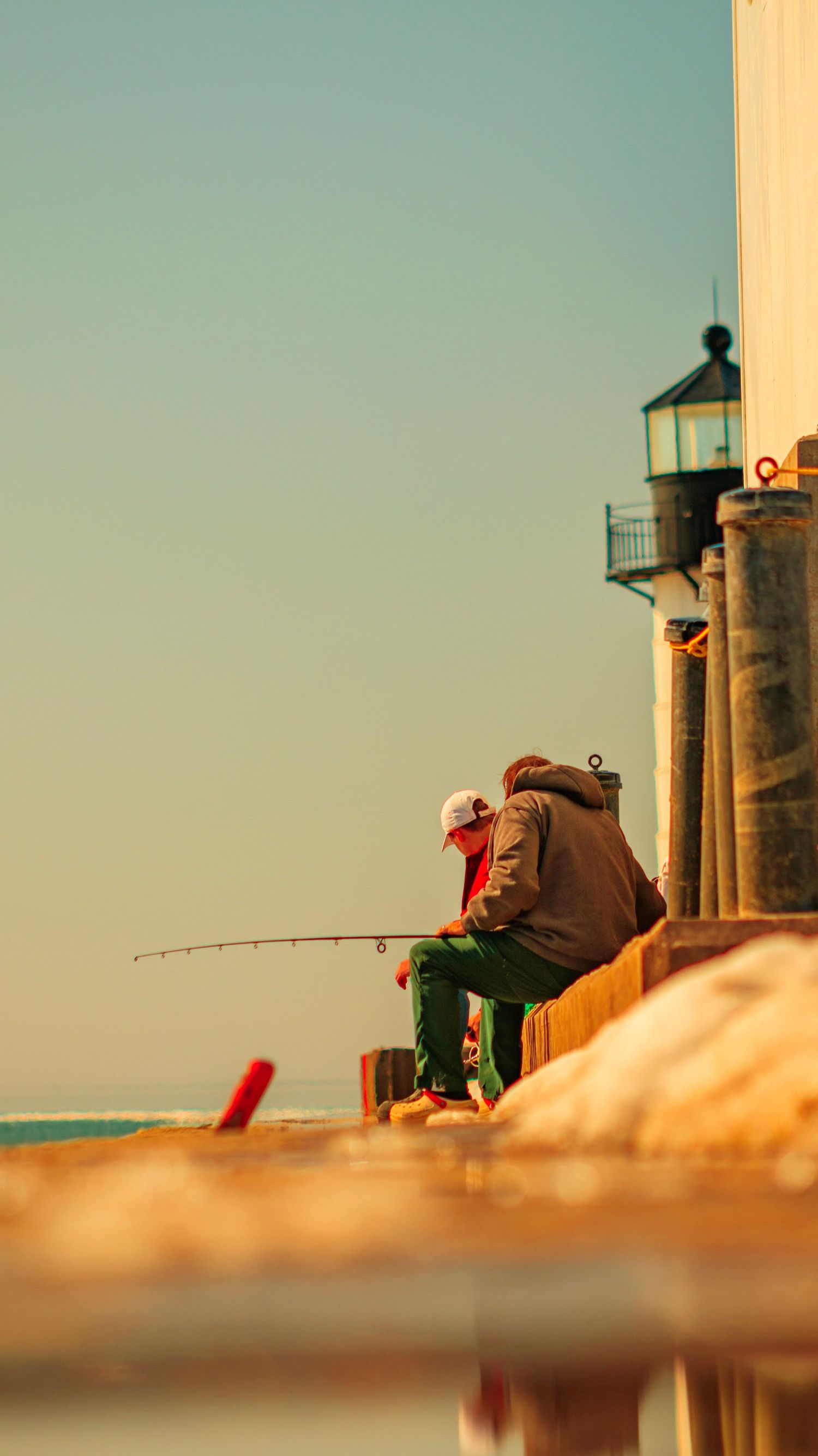 Silhouette of someone fishing near a lighthouse during golden hour.