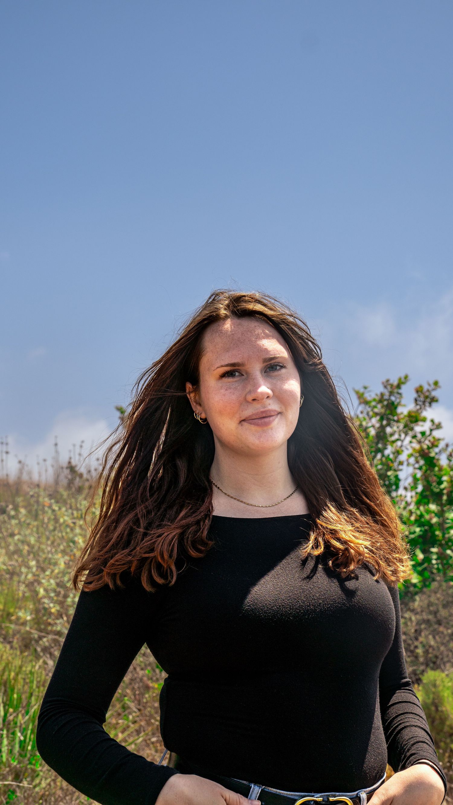 Person in fitted black top and jeans standing in front of sunny hillside with natural vegetation.