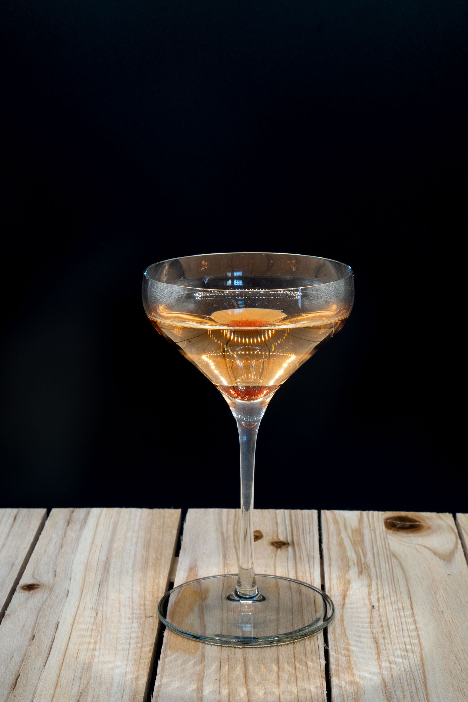 Side view of vintage coupe glass containing amber-colored cocktail on wooden table with dark backdrop.