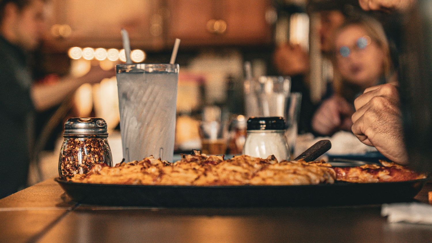 A tall milkshake sits beside a large plate of loaded nachos on a rustic wooden table in a dimly lit restaurant setting.