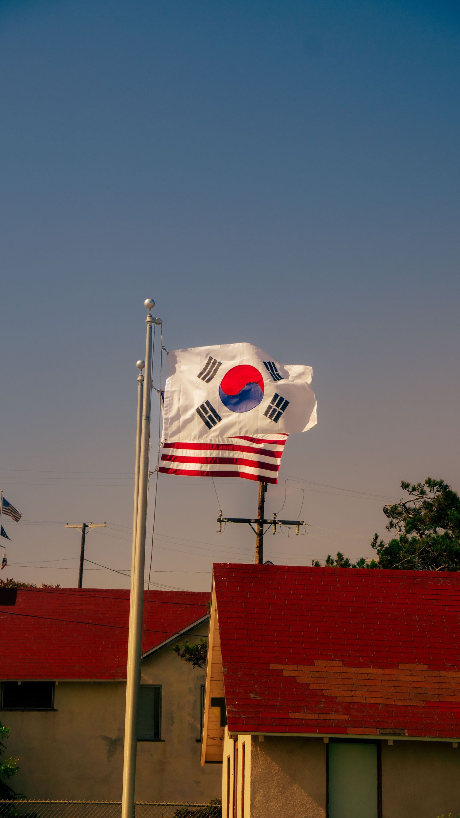 South Korean and American flags waving on pole against blue sky with red roofed buildings below.