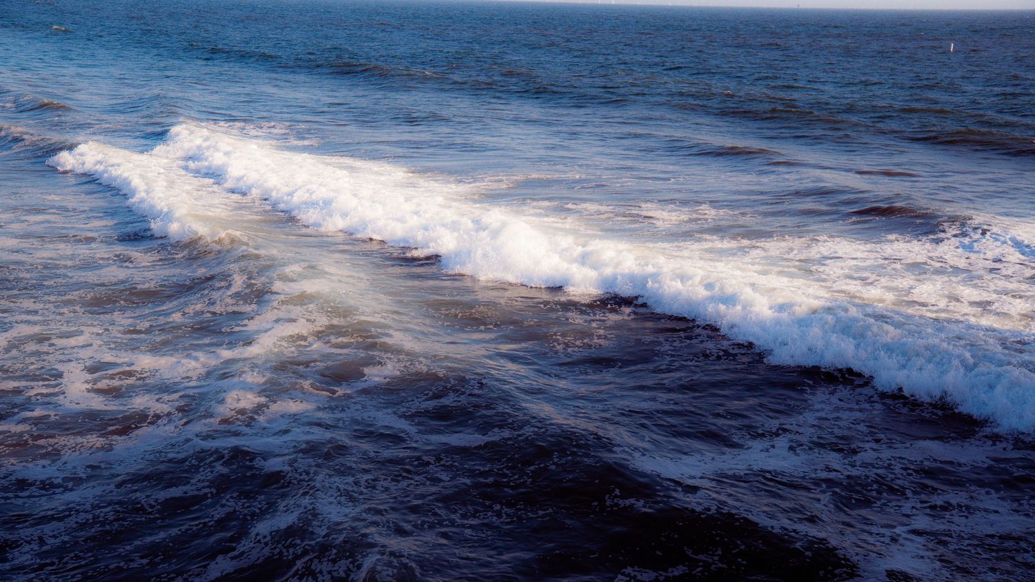 Panoramic seascape showing waves breaking along a dark rocky shoreline.