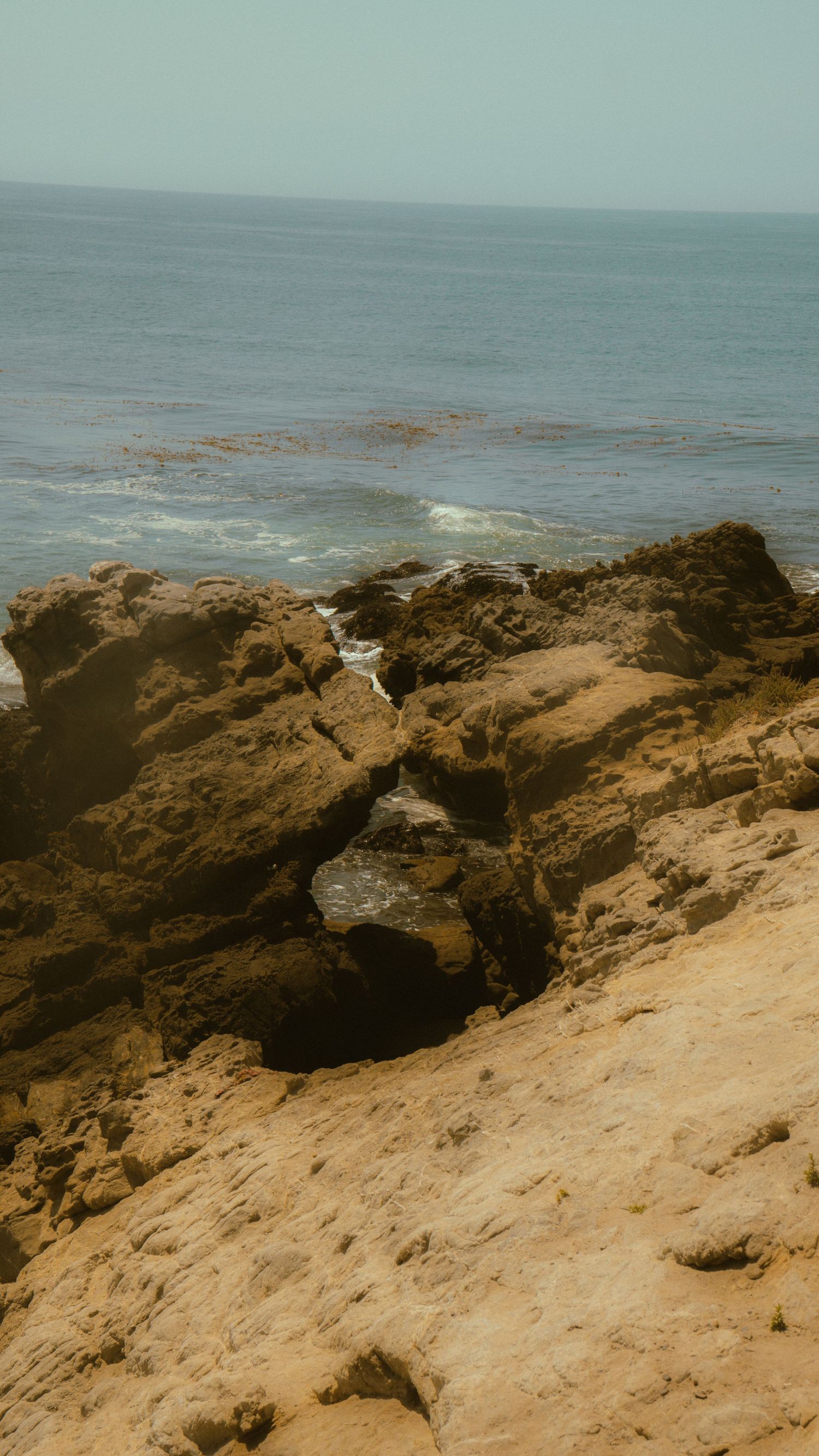 Rugged rocky cliff formation overlooking ocean waves with sandy terrain in foreground.