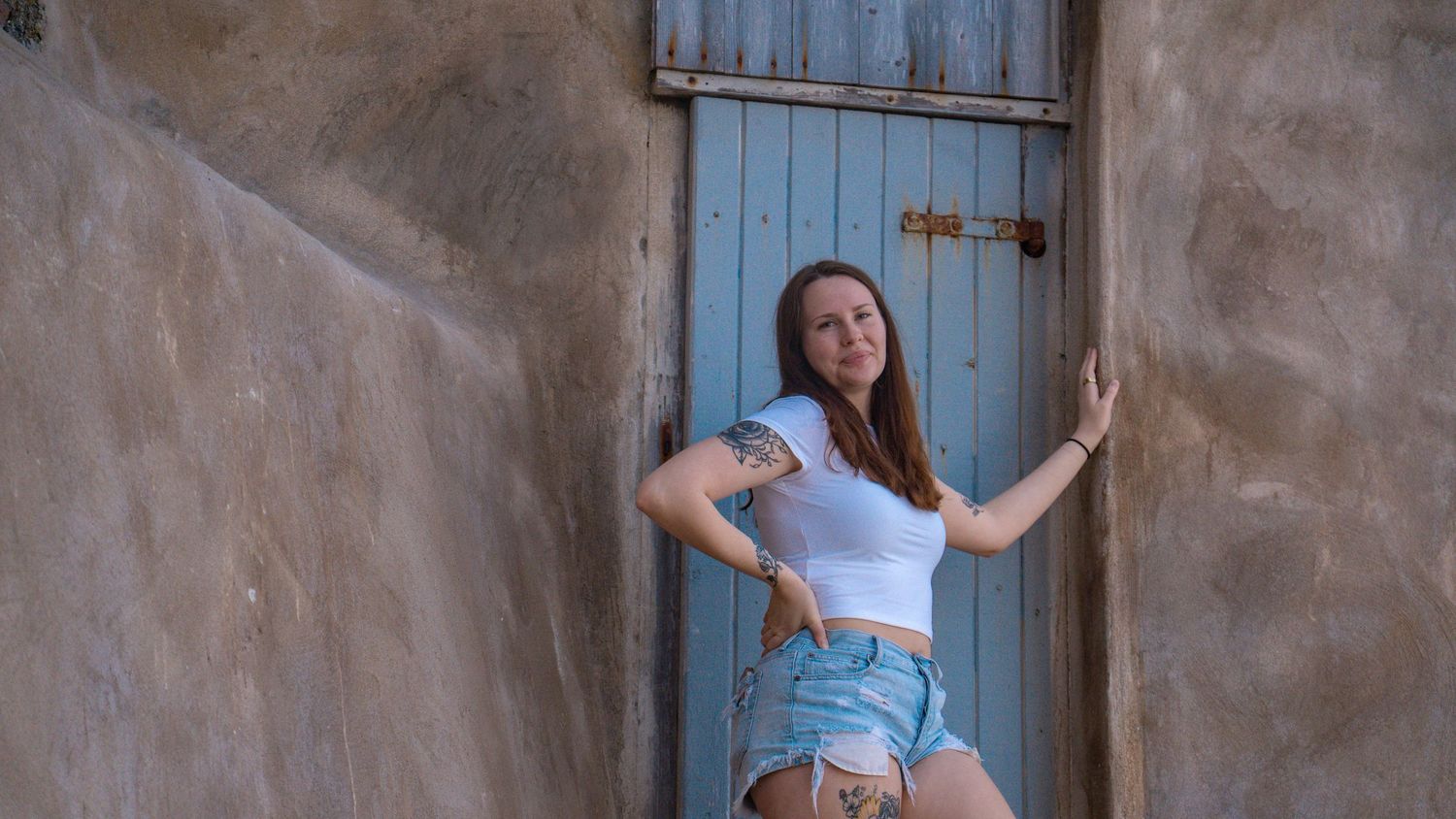 Model poses against rustic blue door in summer outfit showing artistic leg tattoos.