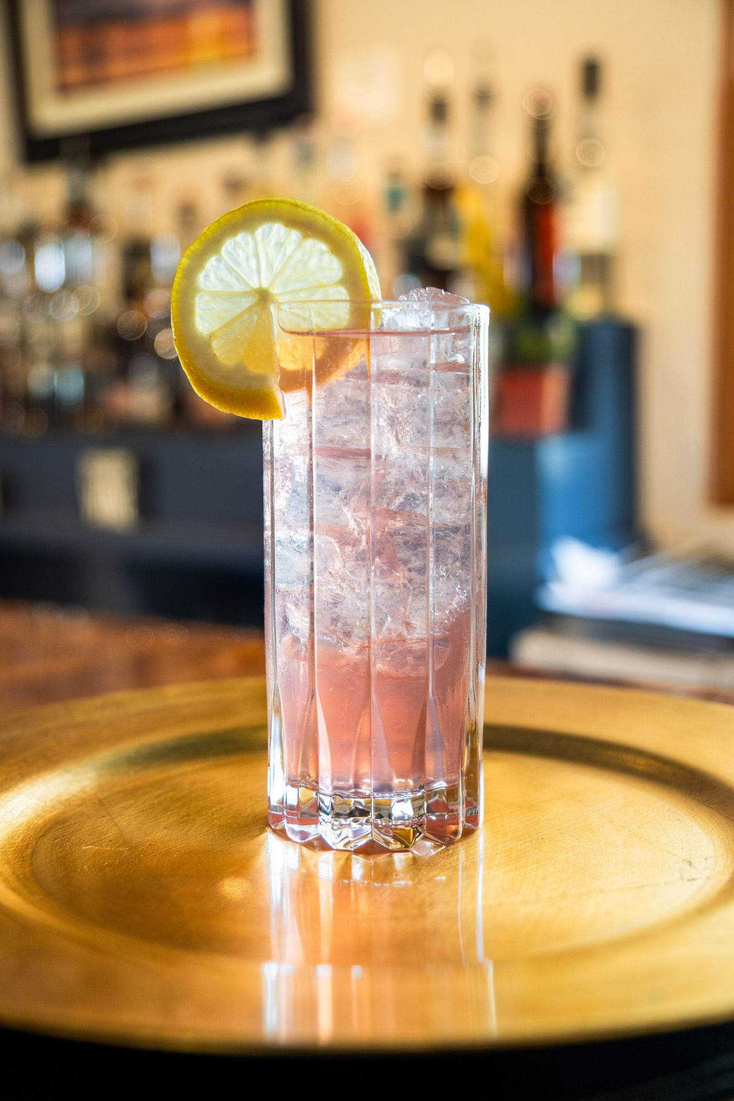 Pink cocktail with lemon wheel garnish served on golden plate with crushed ice.