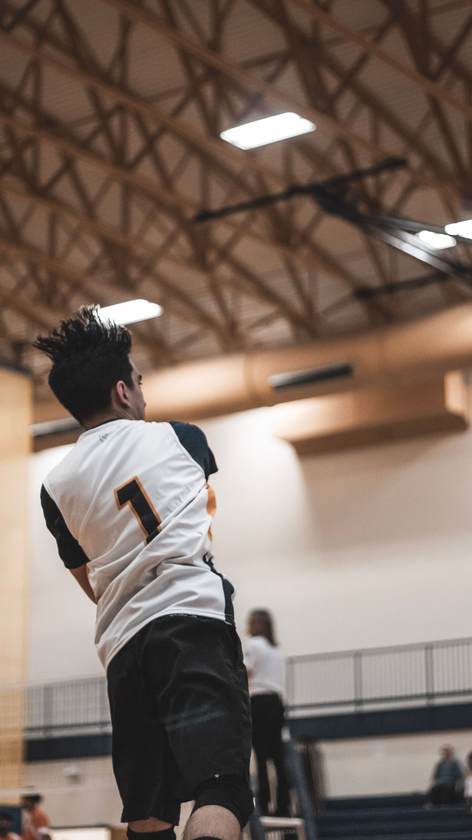 Player in white jersey number 1 jumps to serve volleyball during indoor game.