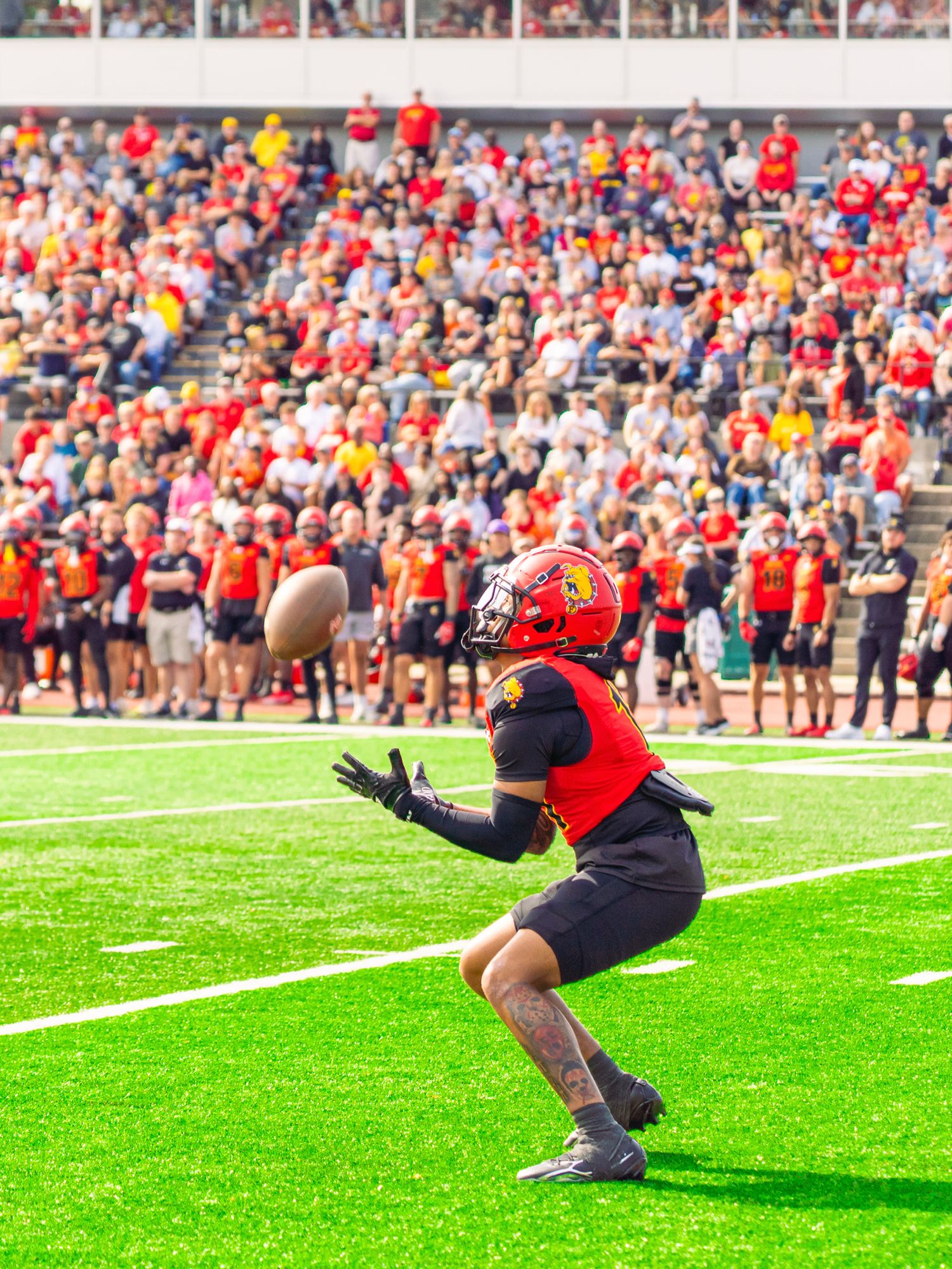 A football player in a red helmet and jersey crouches on turf with hands out, ready to catch a ball as fans cheer in the stands.