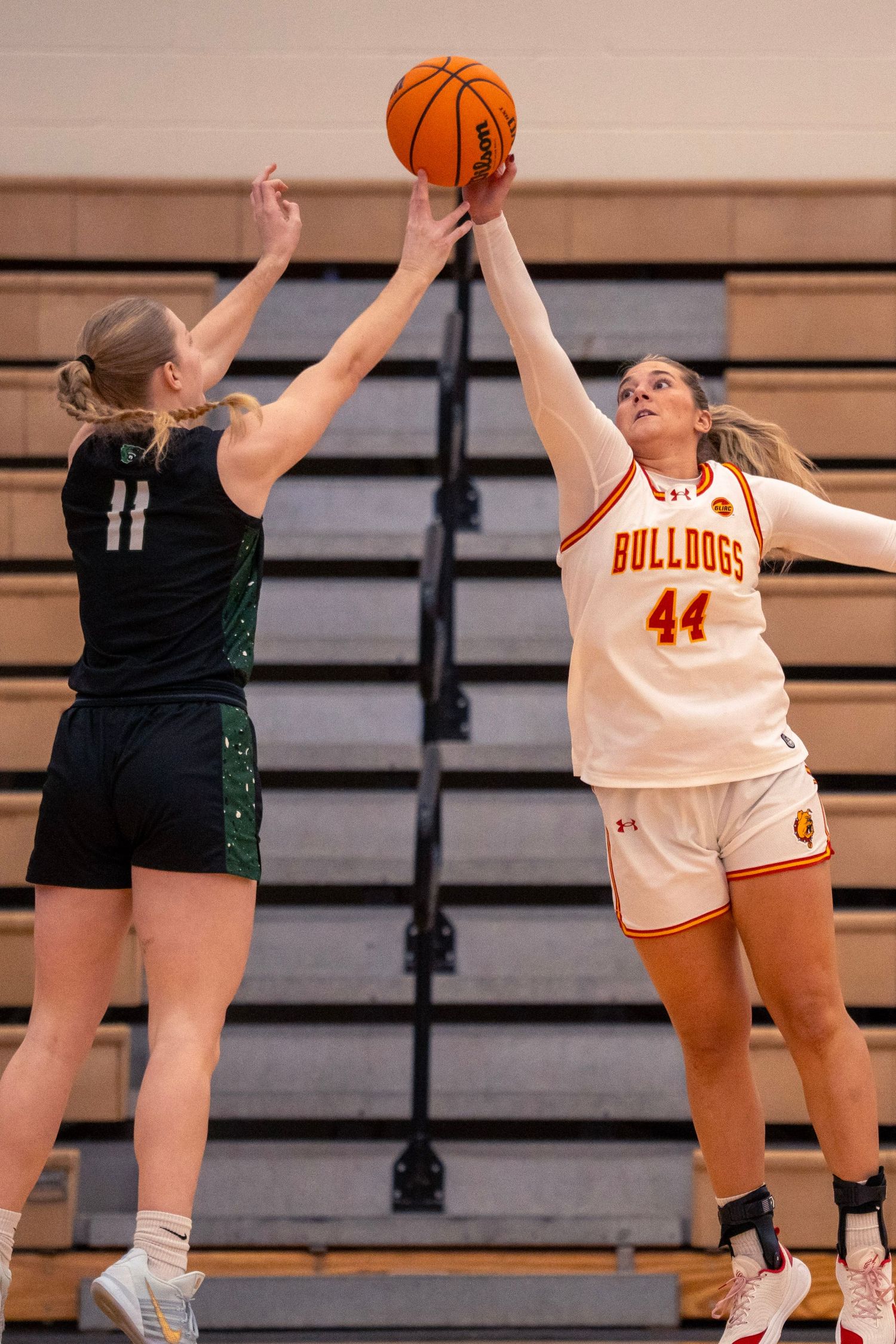 Two women's basketball players leap to contest a shot, one in a white Bulldogs number 44 jersey and one in a dark green number 11 uniform.