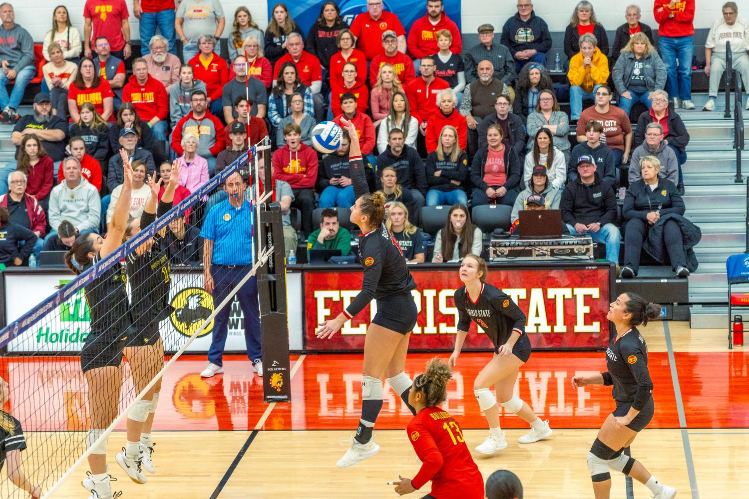 A volleyball player in red uniform jumps to spike the ball during an intense match as spectators watch from the stands.