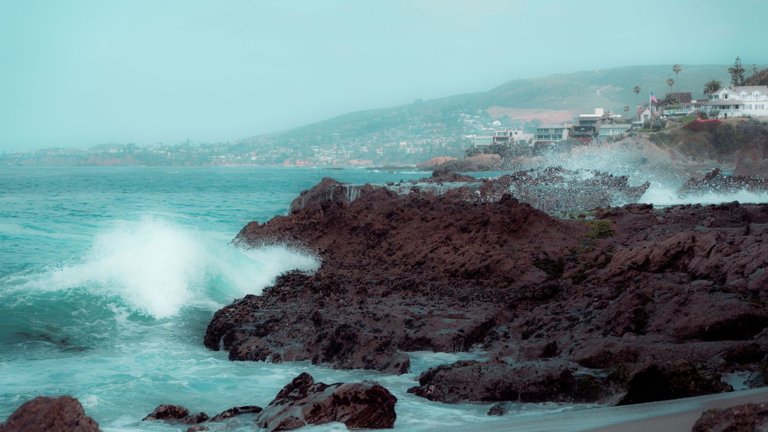 Misty coastal scene with waves breaking against volcanic rocks and distant buildings along the shoreline.