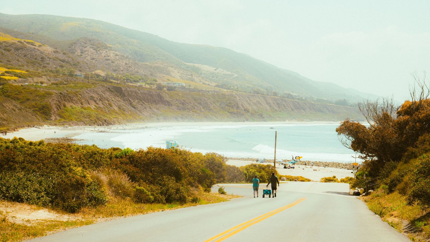 Winding coastal road leads to a pristine beach with mountains in the distance.