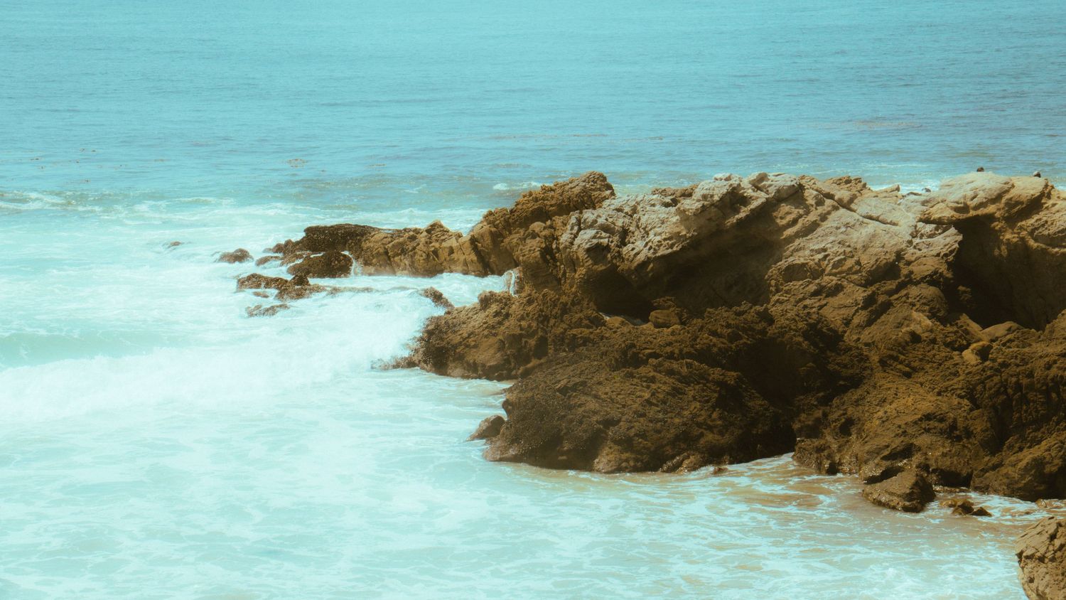 Jagged coastal rocks jutting out into turquoise ocean waters with white foam waves.