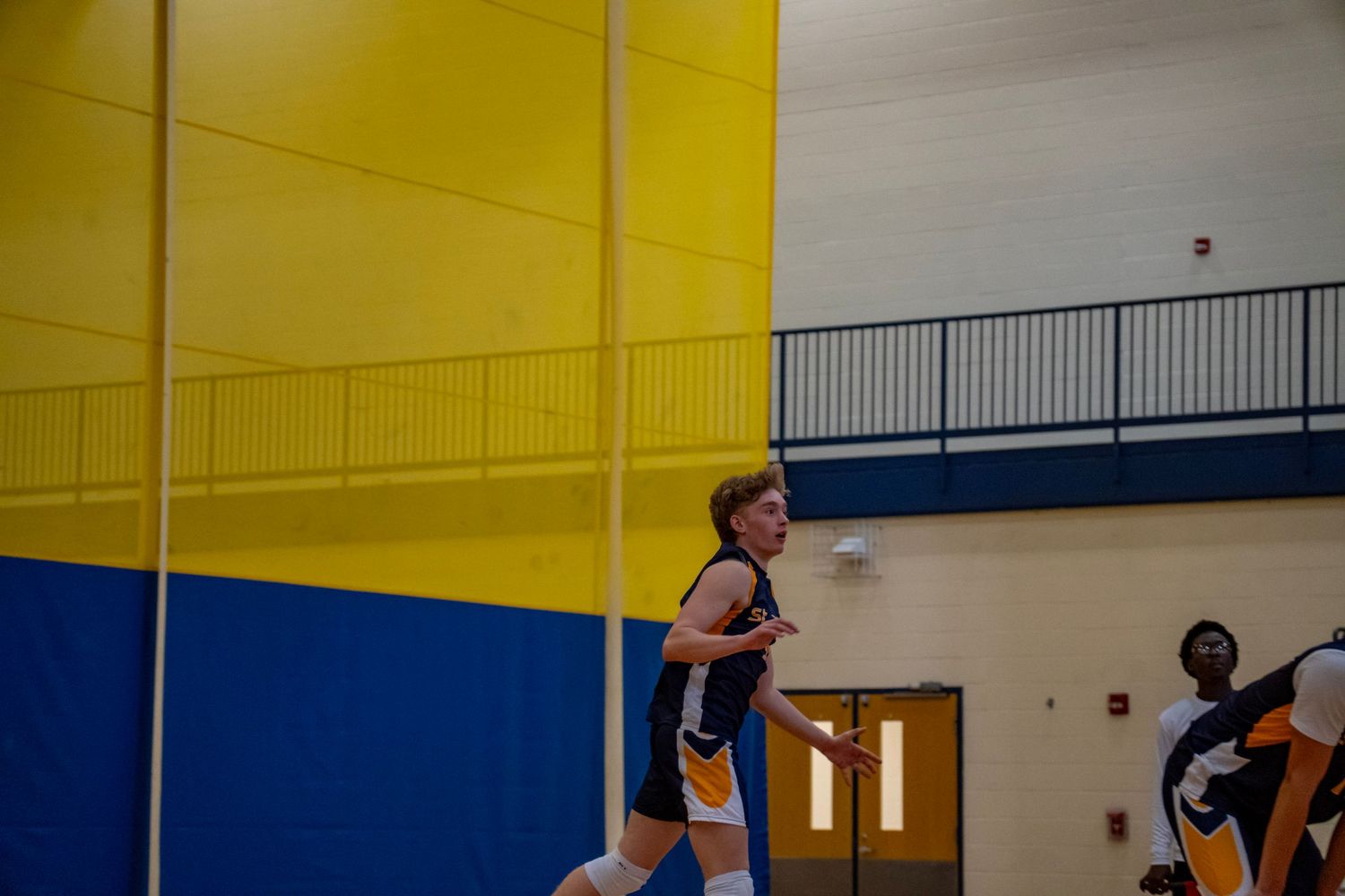 Player jumping to serve in a gymnasium with distinctive yellow and blue walls.