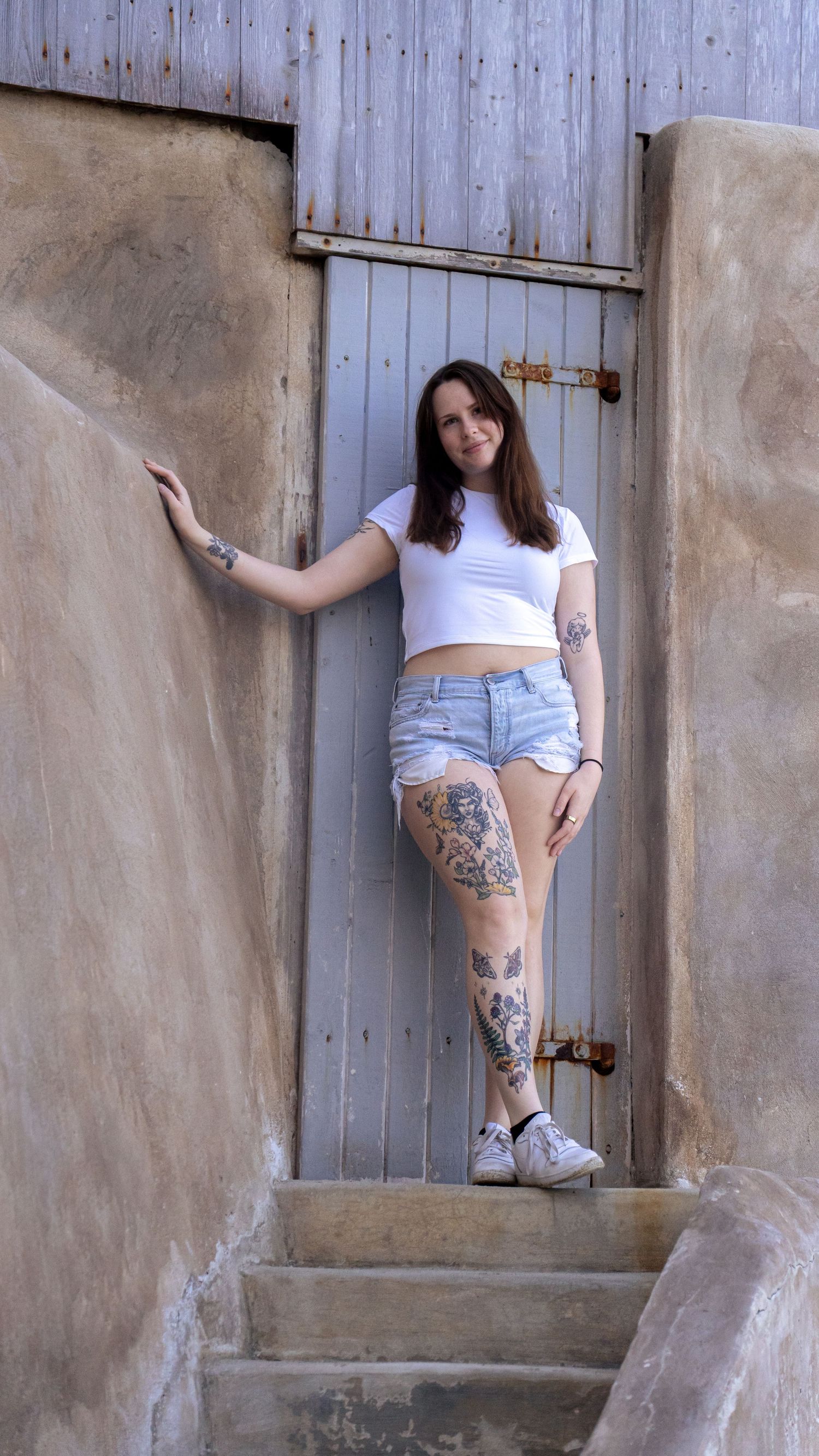 Casual portrait against industrial doorway showing decorative leg tattoos and summer fashion.
