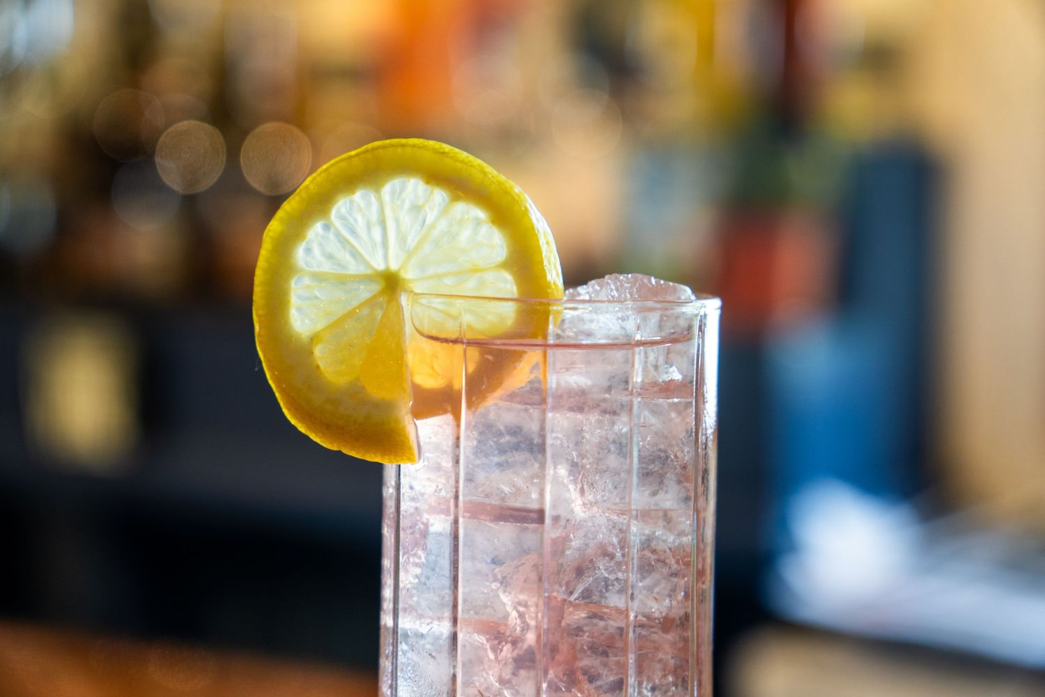 Close-up of pink cocktail with lemon wheel garnish against blurred bar background.