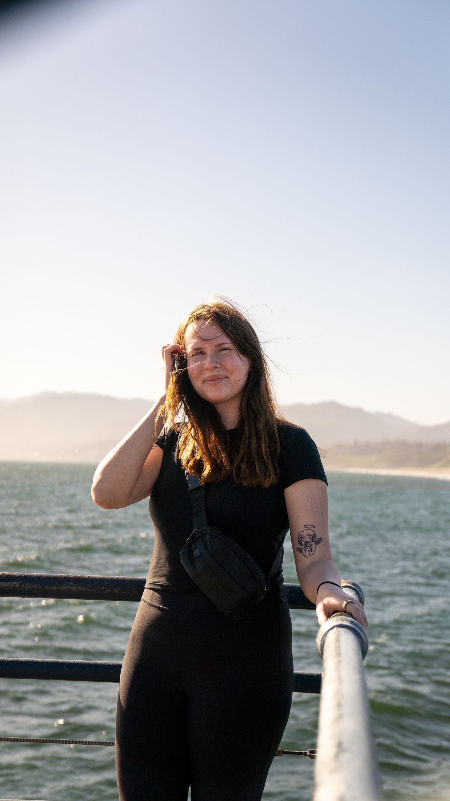 Person wearing black stands by railing overlooking ocean and mountains at sunset.