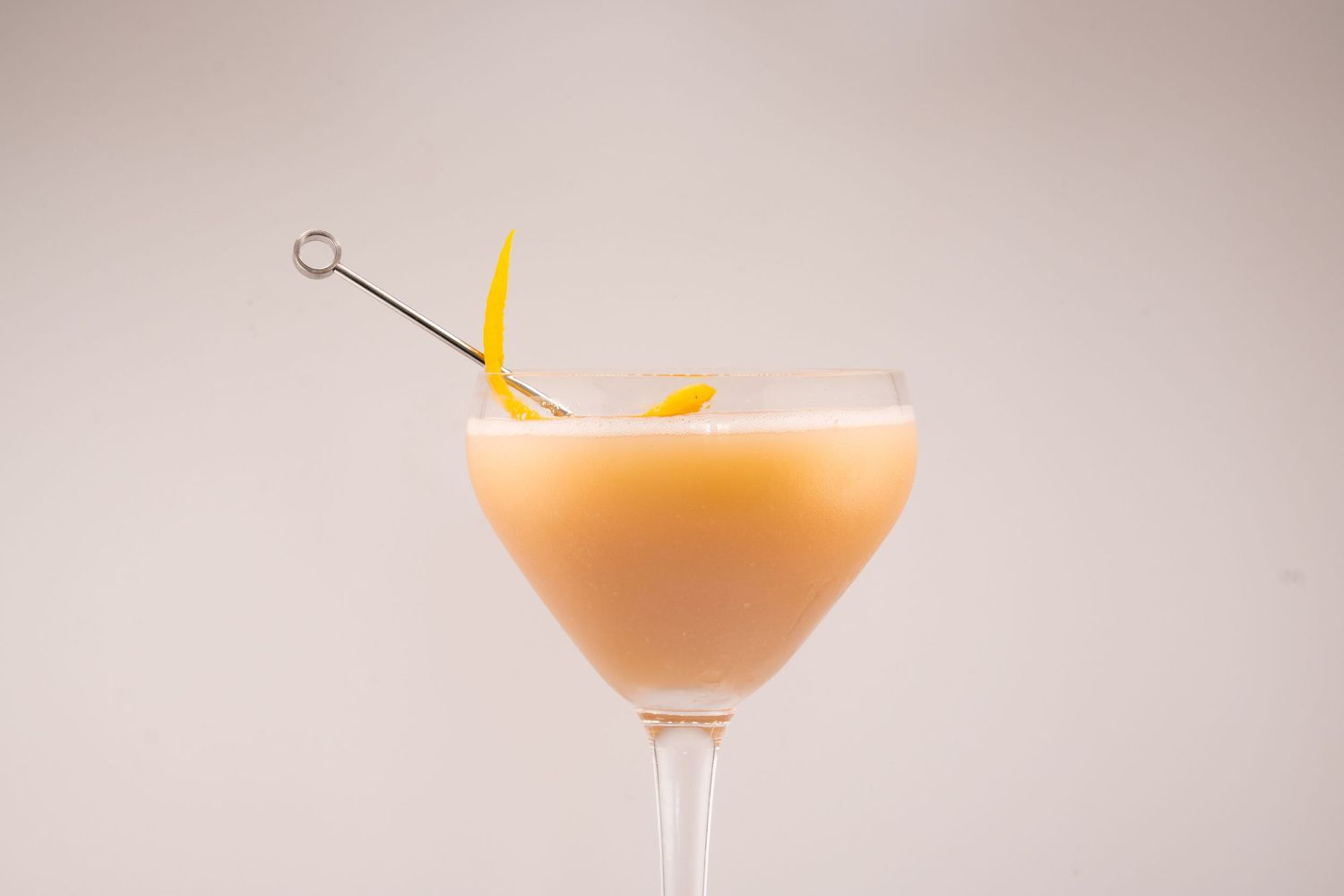 Creamy peach-colored cocktail in martini glass garnished with citrus and metal skewer.