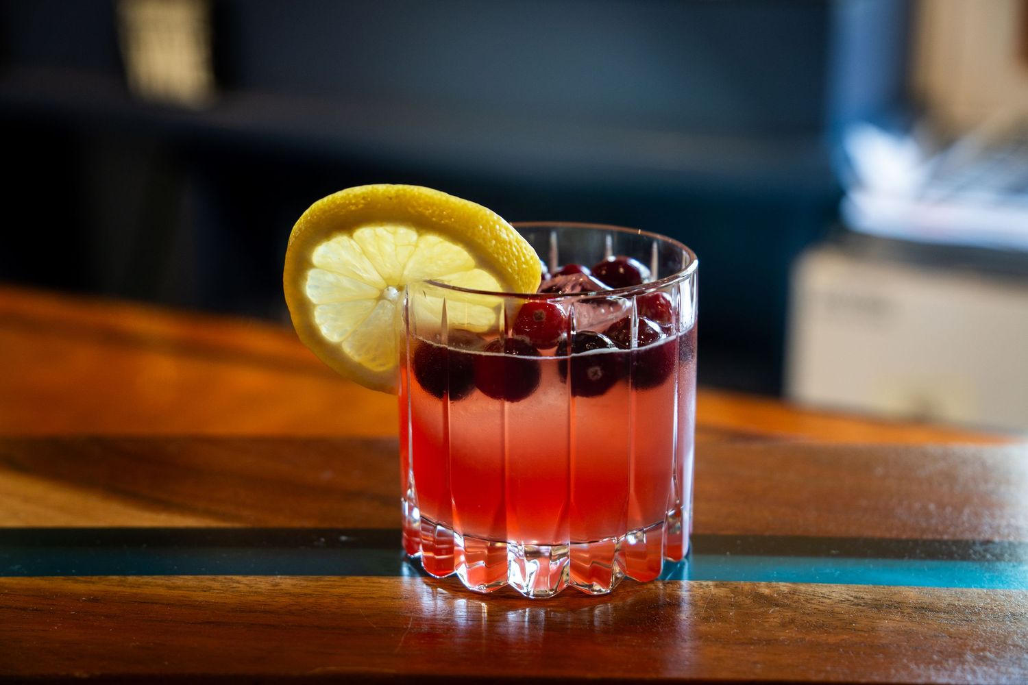 A red cocktail garnished with cranberries and lemon slice served in a rocks glass on a wooden bar.