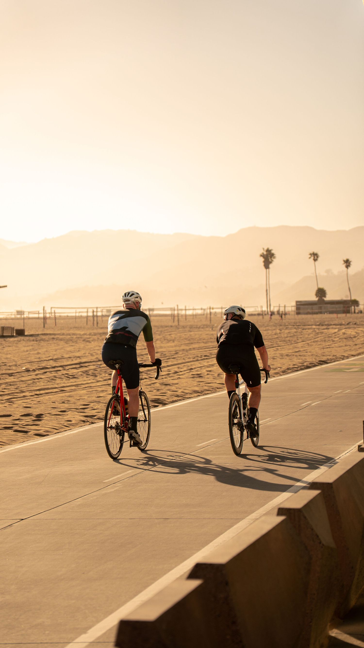 Cyclists riding along concrete path with beach and mountains in sunset background.