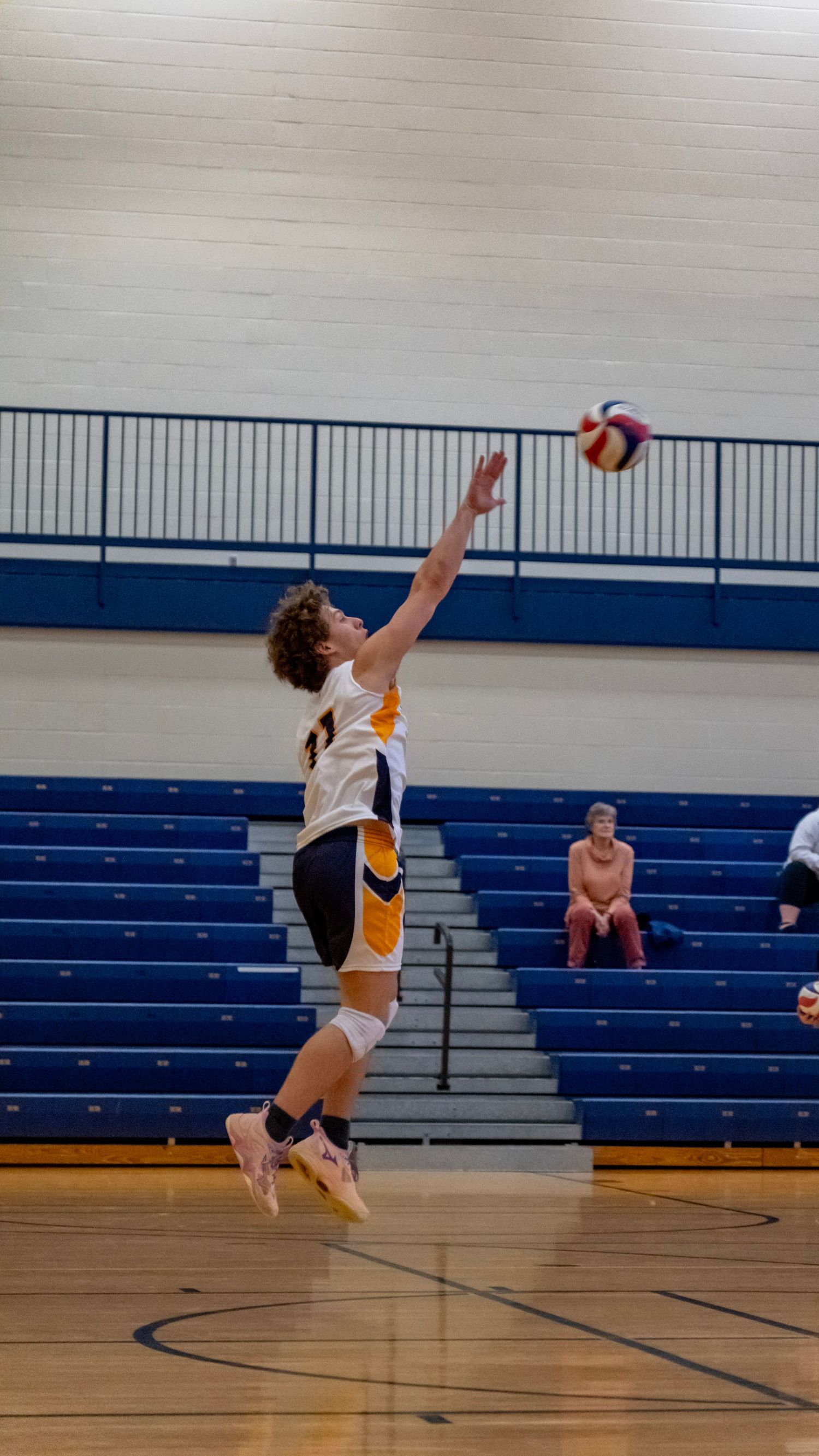 Player jumps high to execute a volleyball serve in a gymnasium with bleachers visible.