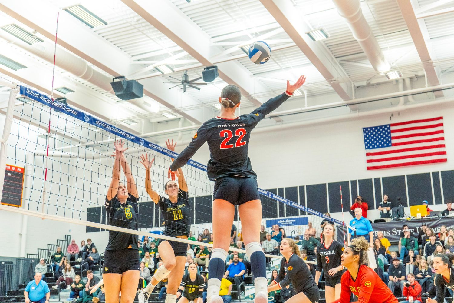 High school volleyball players jump to block a spike during an intense match with American flag visible in background.