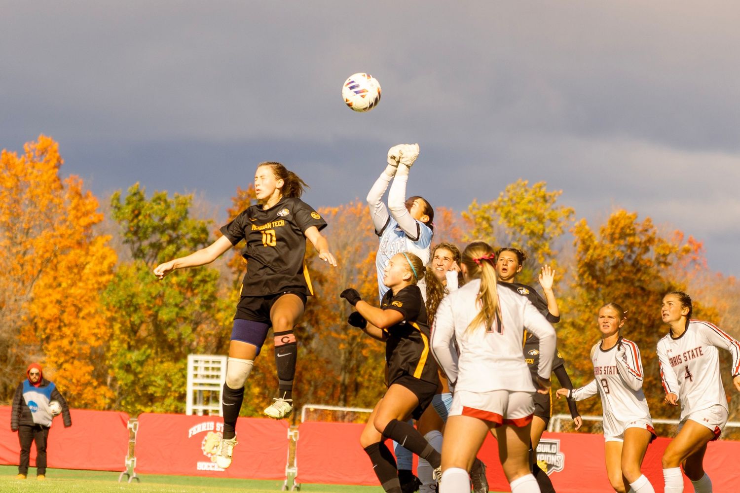 Soccer players leap and compete for a high ball during an autumn match with colorful fall foliage in the background.