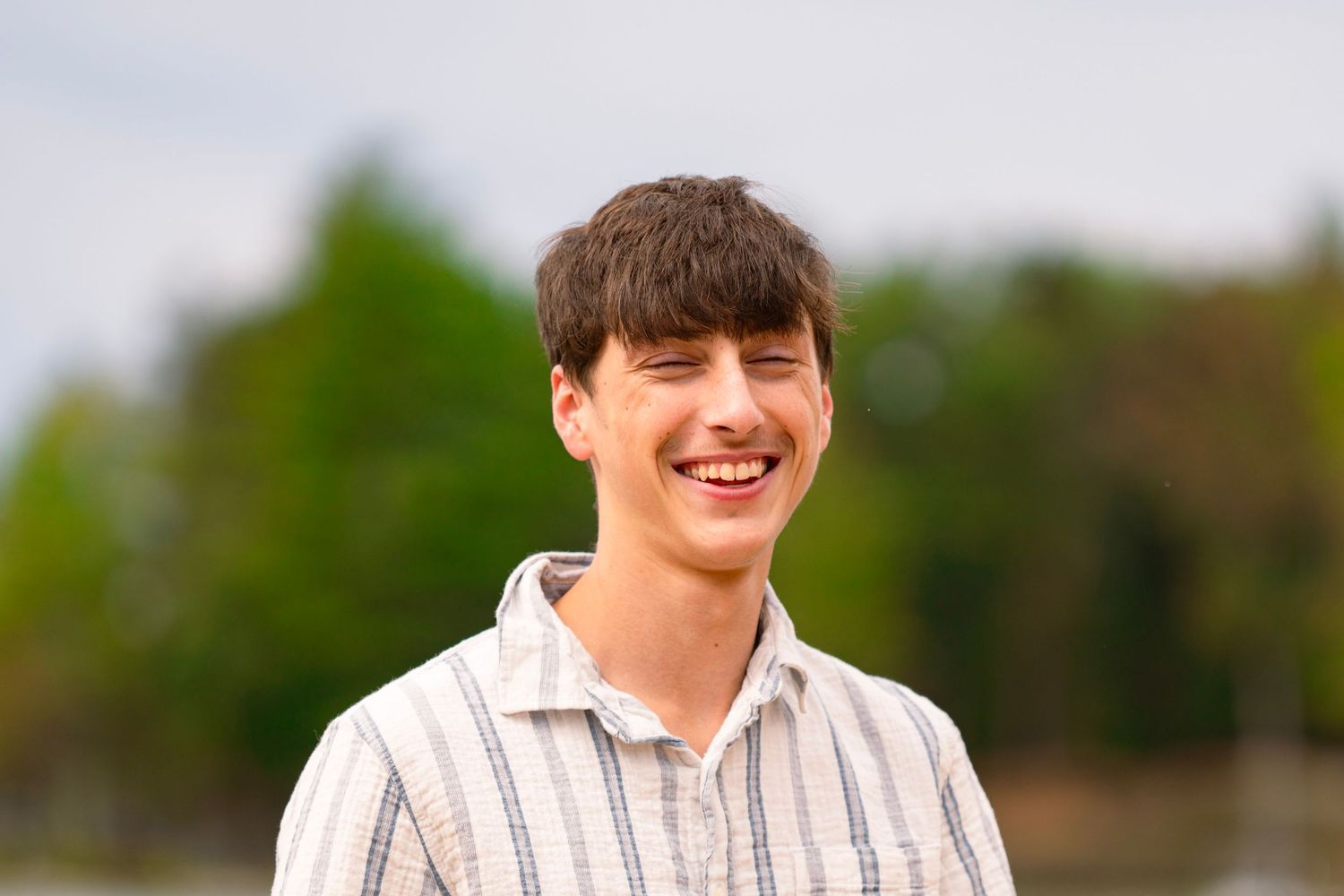Smiling young man with short brown hair wearing a striped button-up shirt, standing outdoors with blurred green trees in the background.