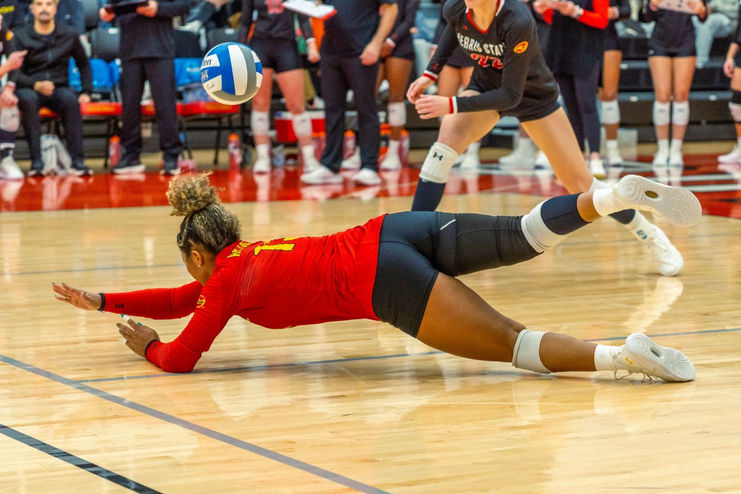 Volleyball players dive and stretch across the court floor during an intense match sequence.