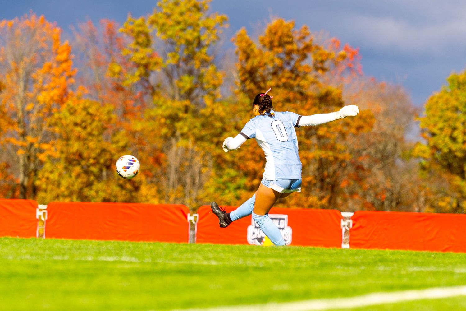 Soccer players celebrate with raised arms on a sunny autumn field with vibrant fall foliage in the background.