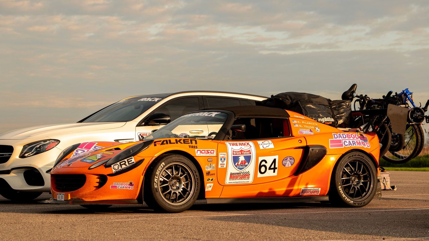Orange race car with number 64 and white Mercedes parked next to it at sunset.