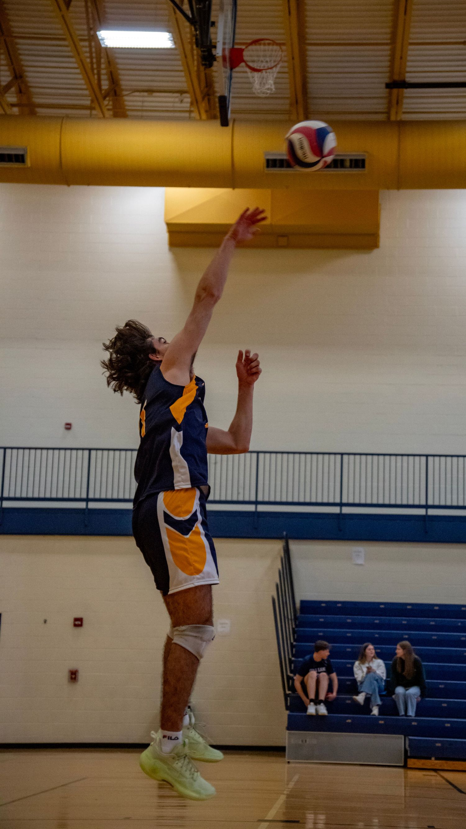 Player in navy and gold uniform jumps to serve the volleyball in an indoor gymnasium.
