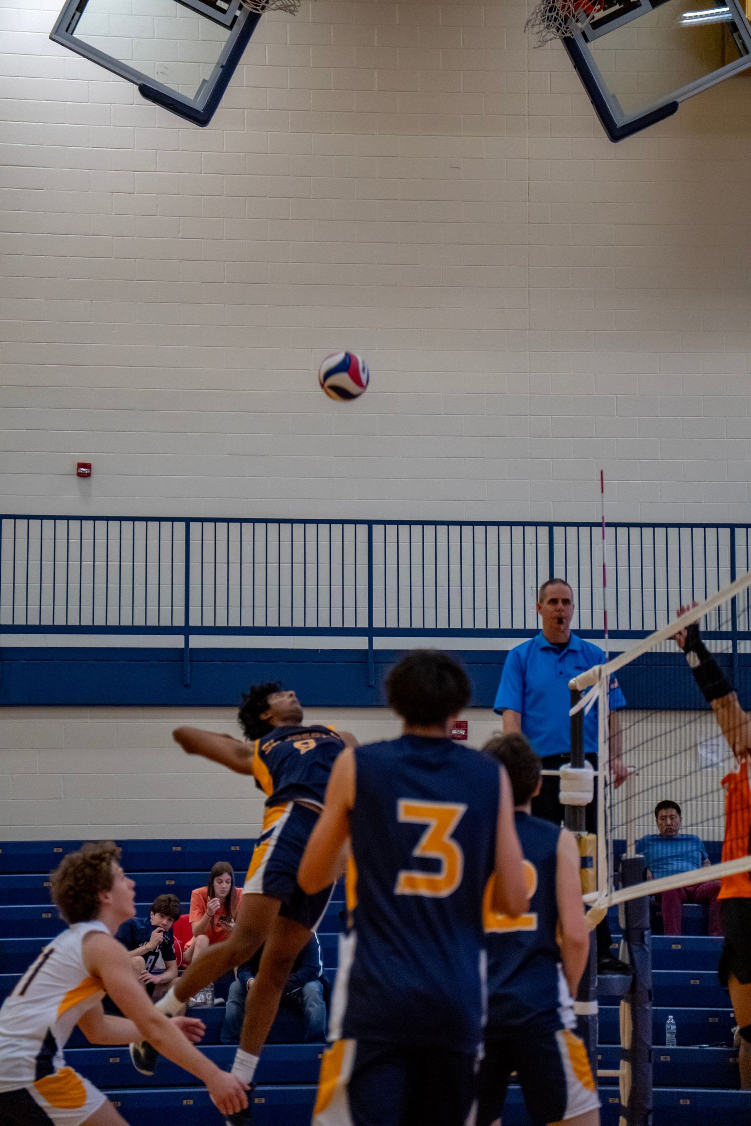 Indoor volleyball match showing players positioning as the ball floats above.